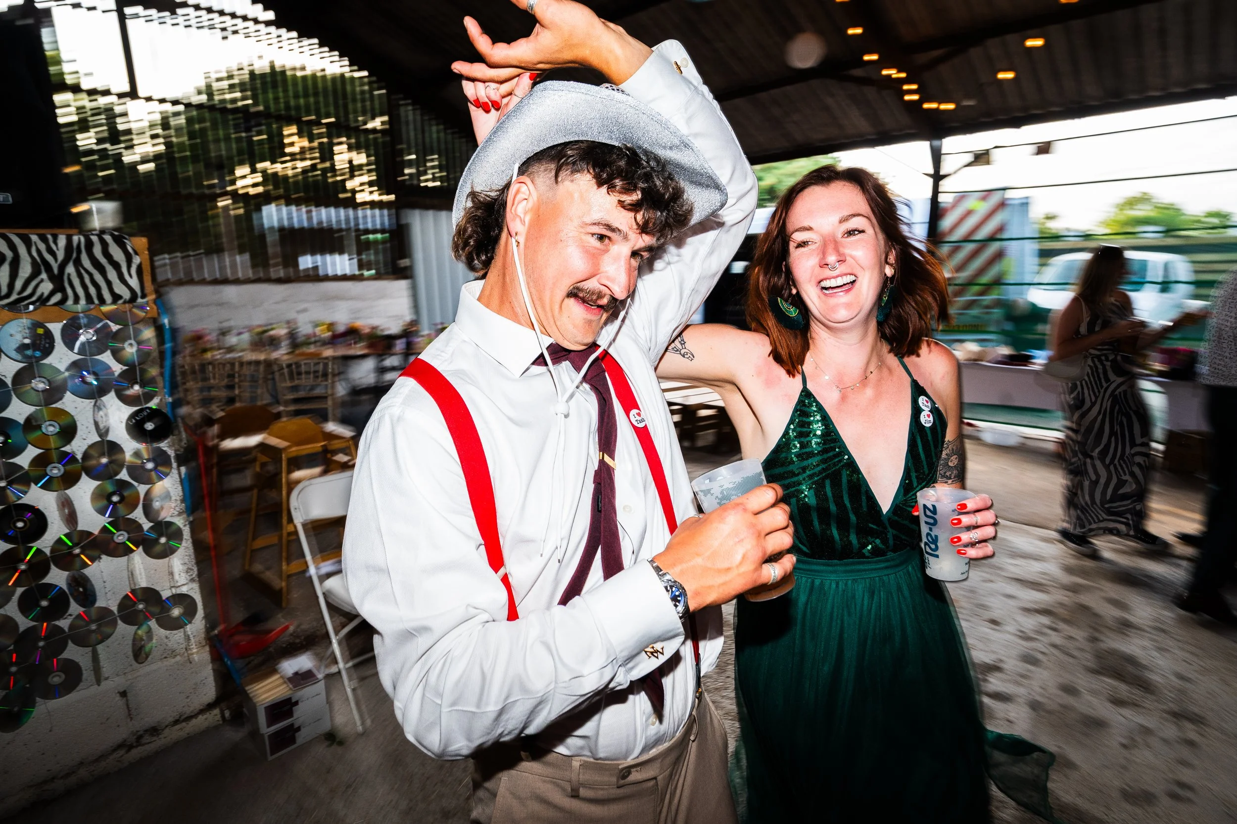 A man and woman laughing and enjoying drinks at an indoor event. The man is wearing a gray hat, white shirt, red suspenders, and a tie, while the woman has shoulder-length hair, green dress, and earrings. They are holding plastic cups, and there are 