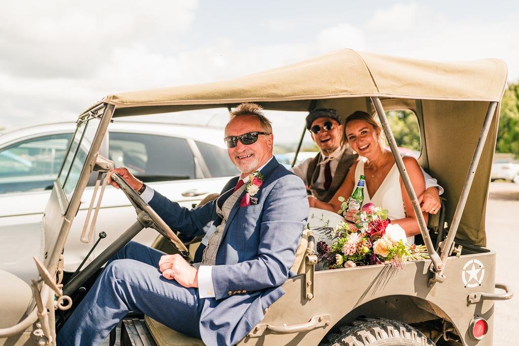 Three people in a vintage jeep with a beige canvas roof, dressed for a wedding, smiling and holding flowers.