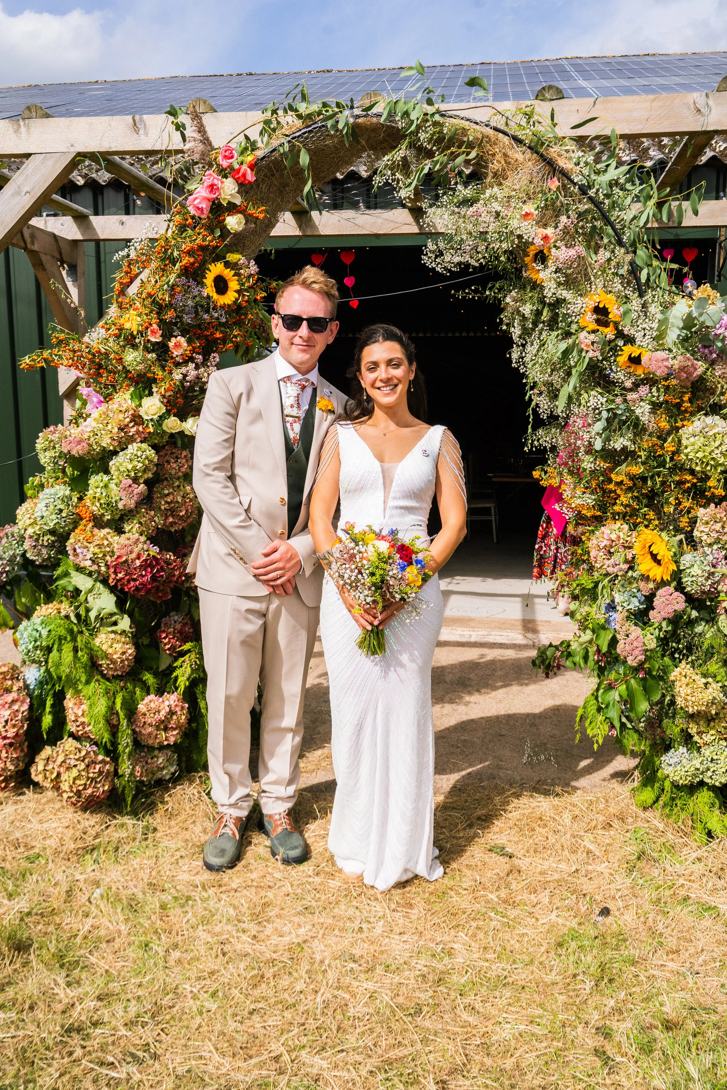 A bride and groom stand under a floral archway at their wedding, smiling for the camera.