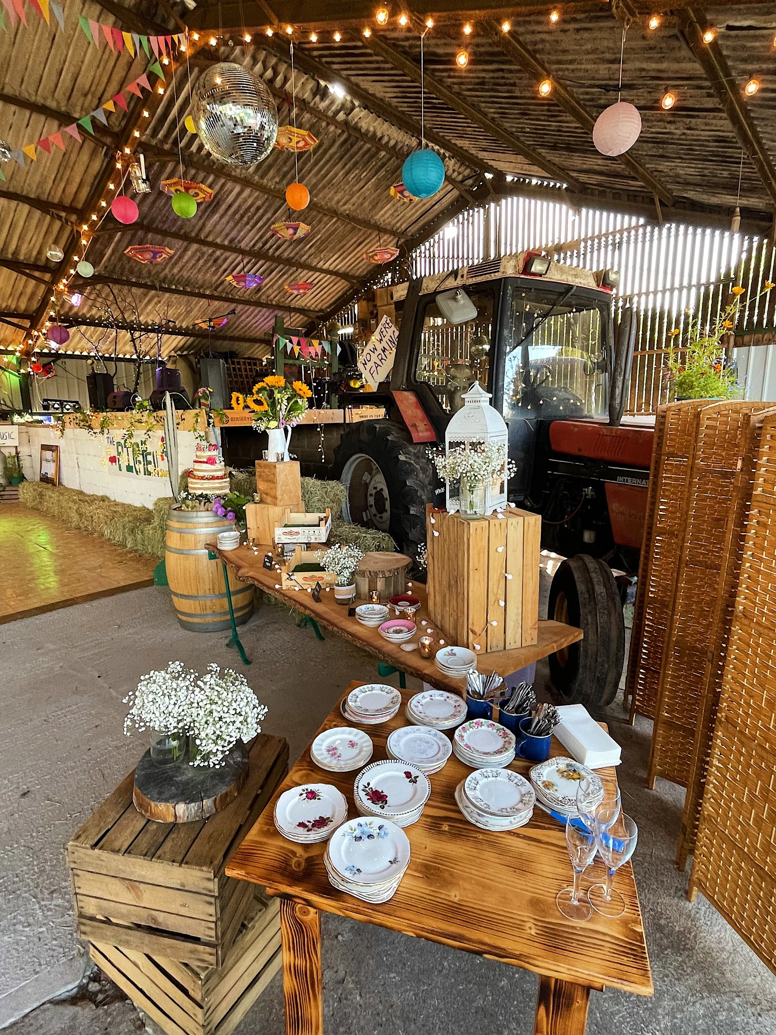 Decorated rustic barn with string lights, paper lanterns, a mirrored disco ball, a tractor, and a table with plates, glasses, flowers, and a cake, suggesting a festive celebration.