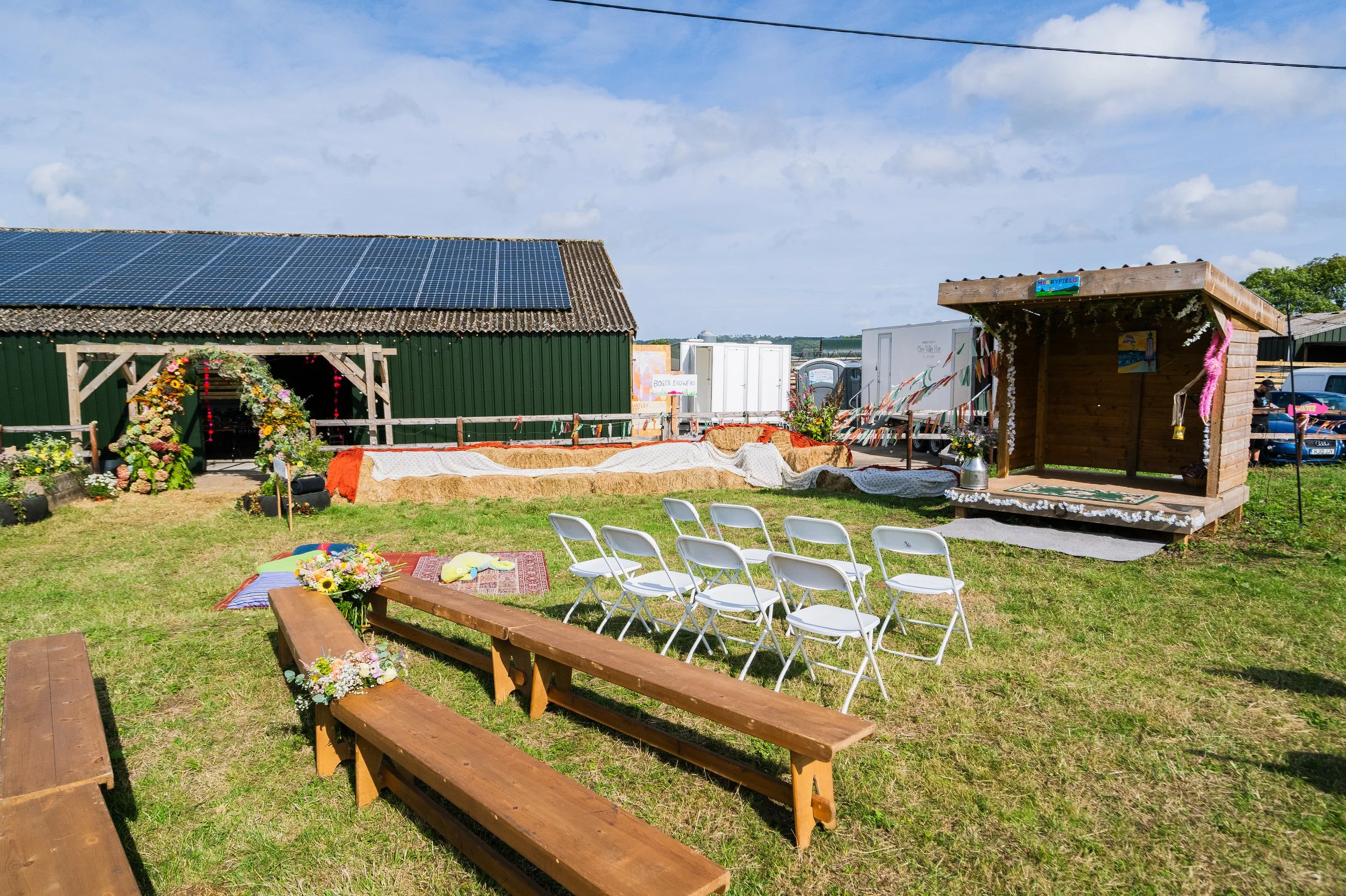 Outdoor wedding setup with white chairs facing a floral arch, wooden benches with flowers, a decorated small wooden stage, and a green building with solar panels under a blue sky.