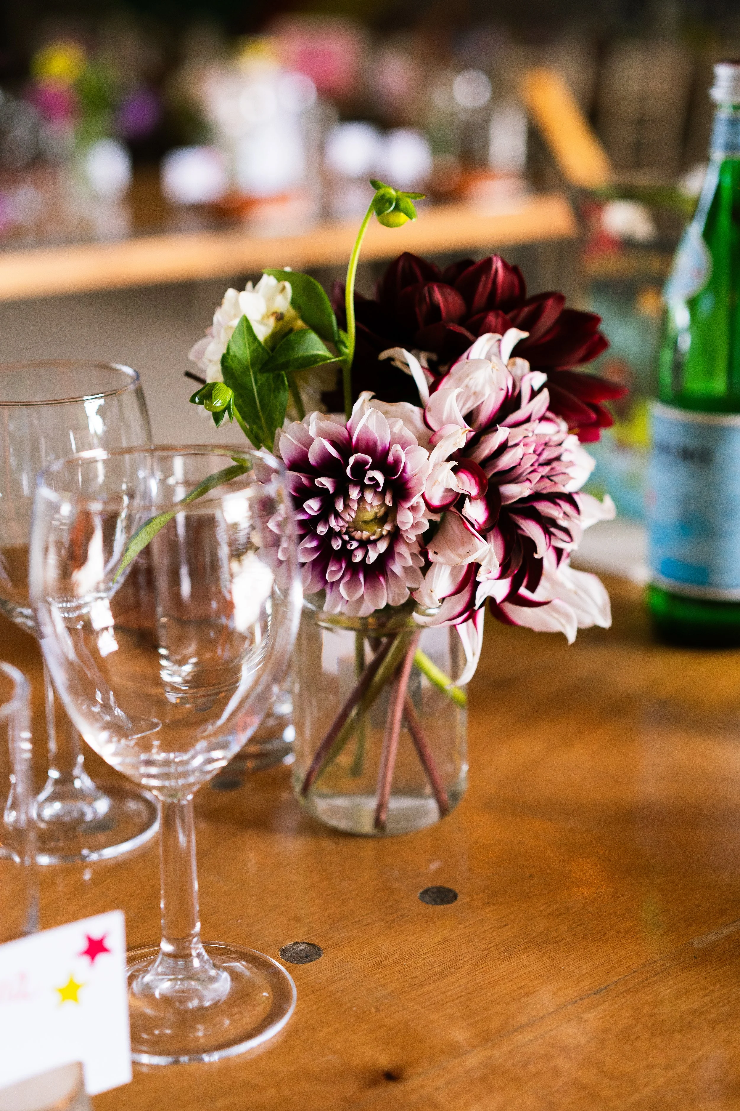 A bouquet of colorful flowers in a glass vase on a wooden table with empty wine glasses and a green bottle in the background.