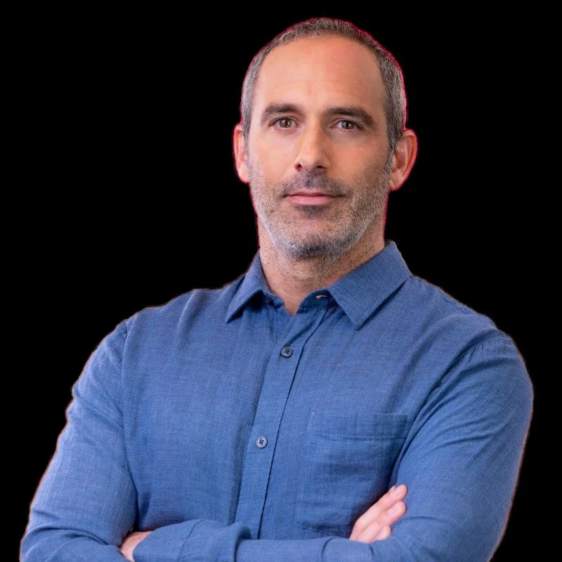 Amer Chehab: a man with short gray hair and a beard, wearing a blue button-up shirt, standing with arms crossed against a black background.