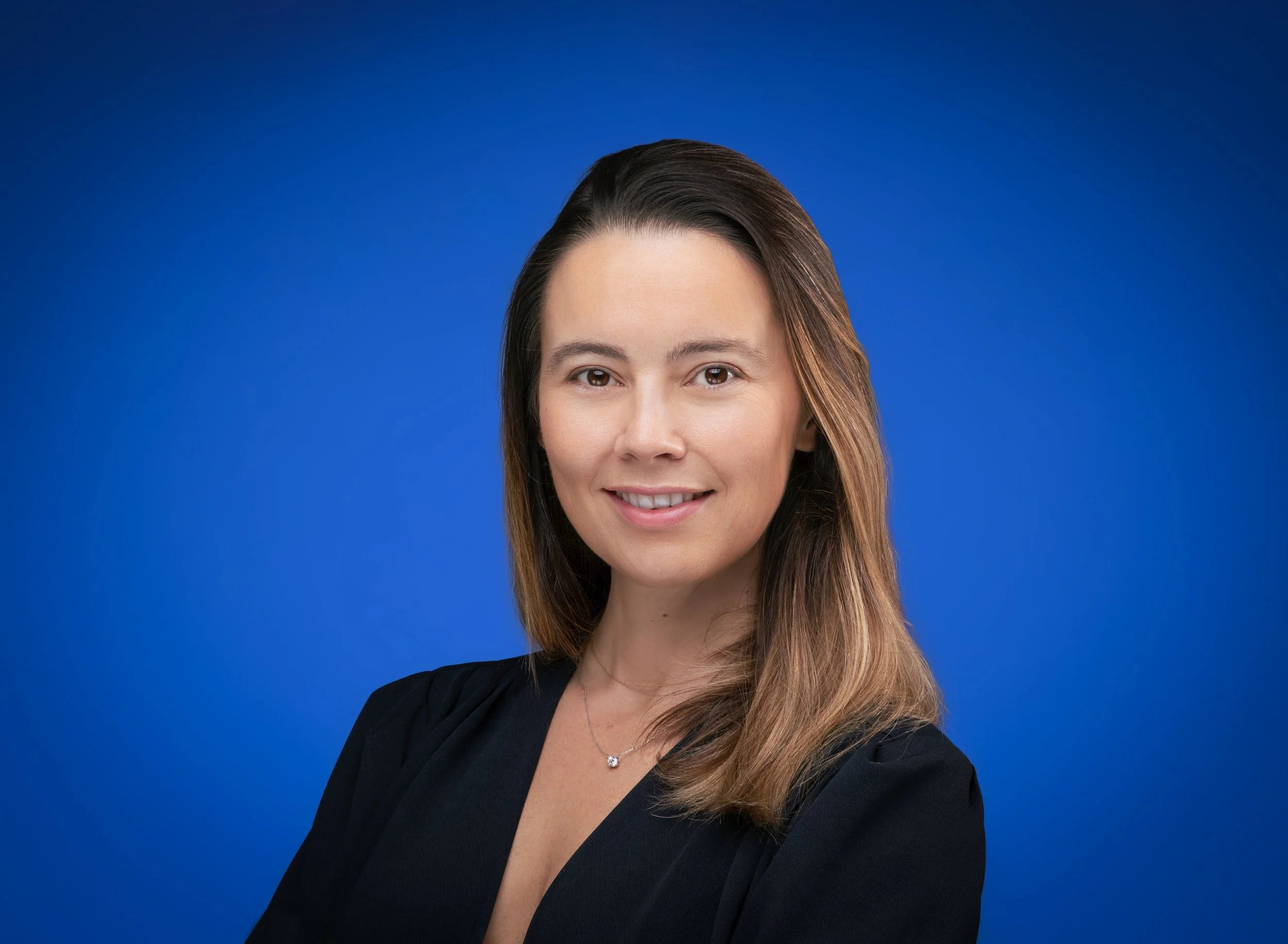 Marie De Ducla: a smiling woman with shoulder-length brown hair, wearing a black top and a delicate necklace, against a blue background.