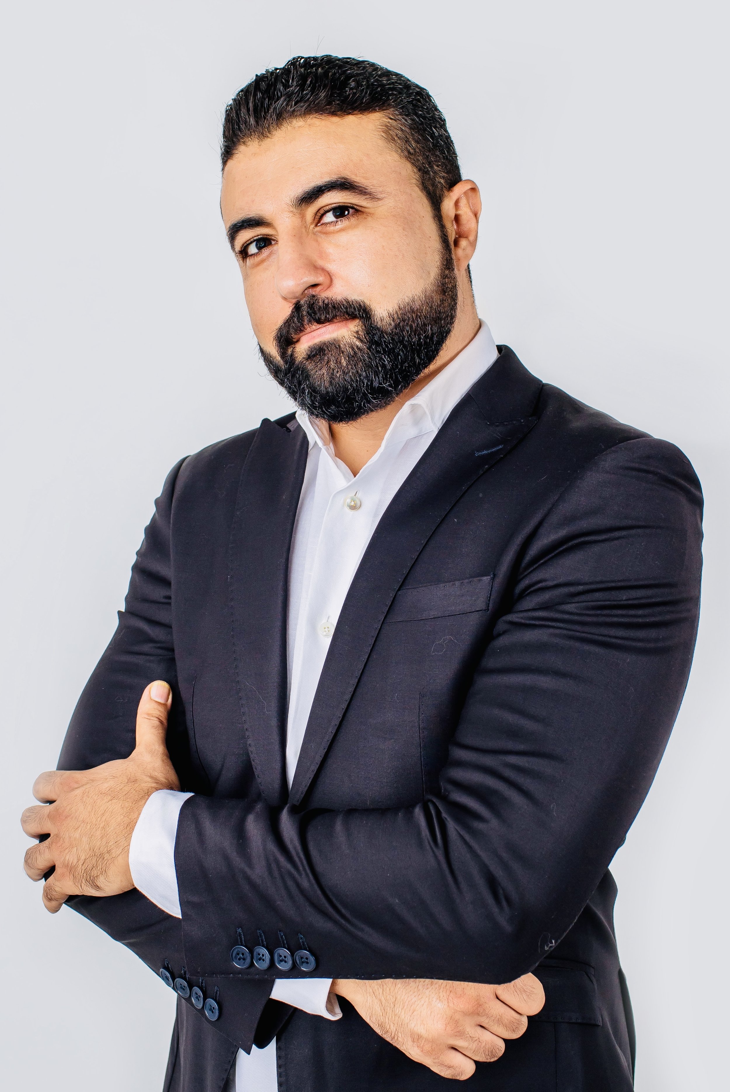 Amine Sadik: a man with dark hair and a beard wearing a black suit and white shirt, standing with arms crossed against a plain light gray background.