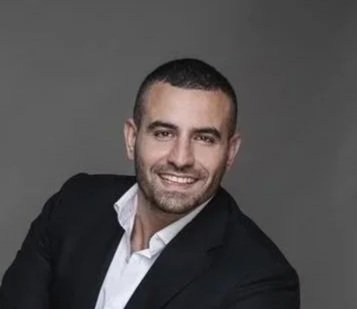 Tony Wazen: a man in a black blazer and white shirt sitting on a black chair against a gray background, smiling with arms crossed.