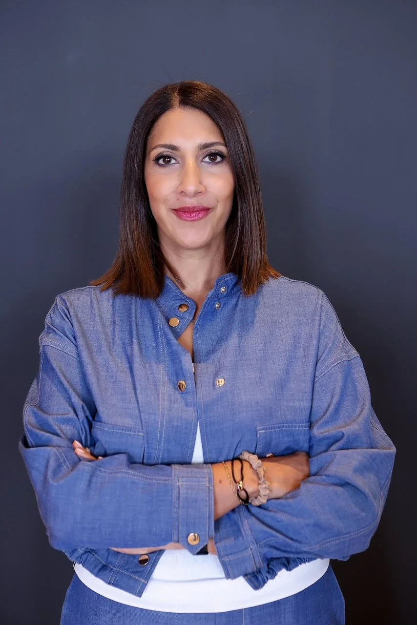 Olfa Messaoudi: a woman with straight brown hair, wearing a denim jacket over a white top, standing against a dark background with arms crossed and a slight smile.
