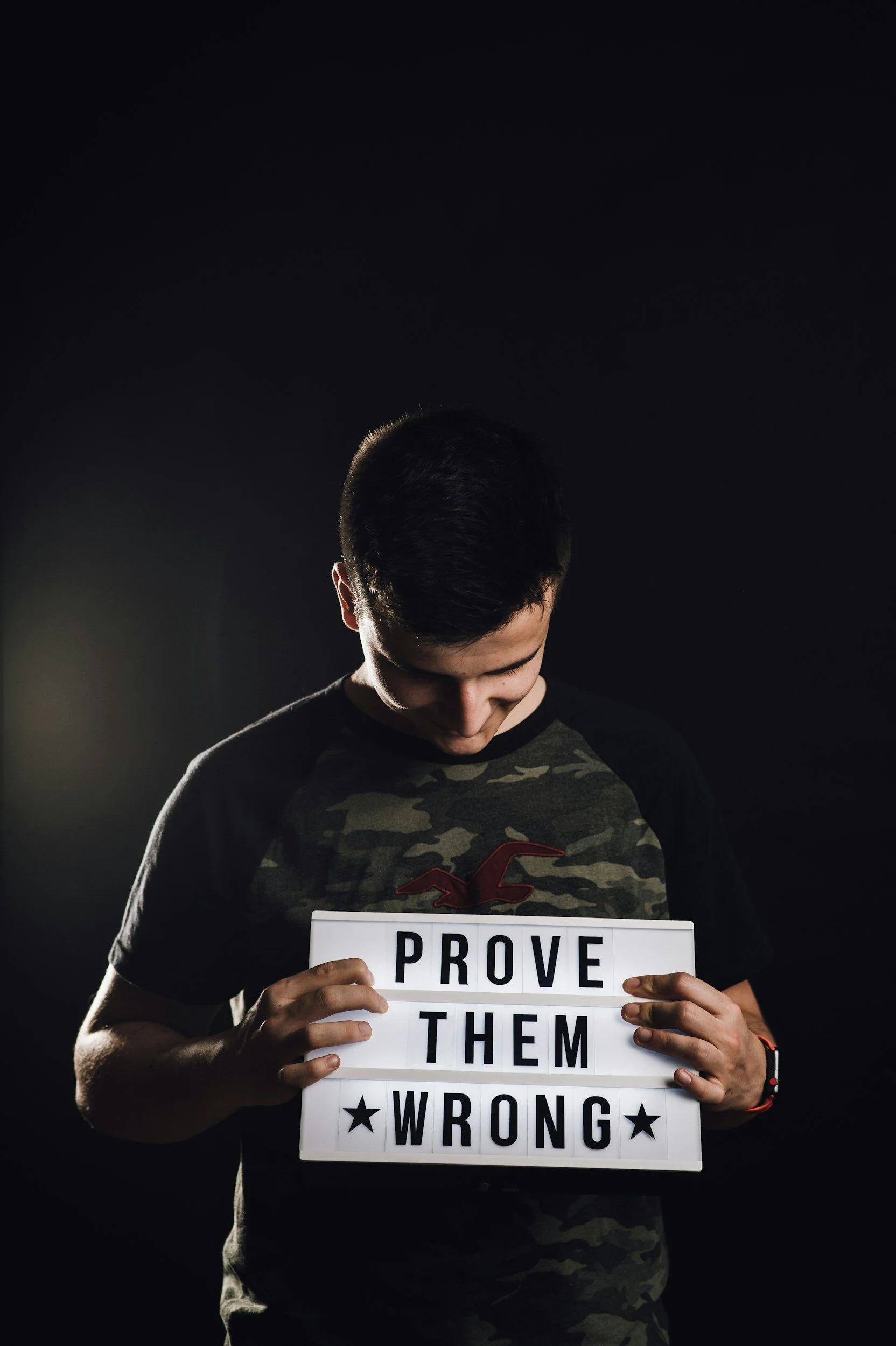 A young man with dark hair looking downward and holding a sign that says 'PROVE THEM WRONG' against a dark background.