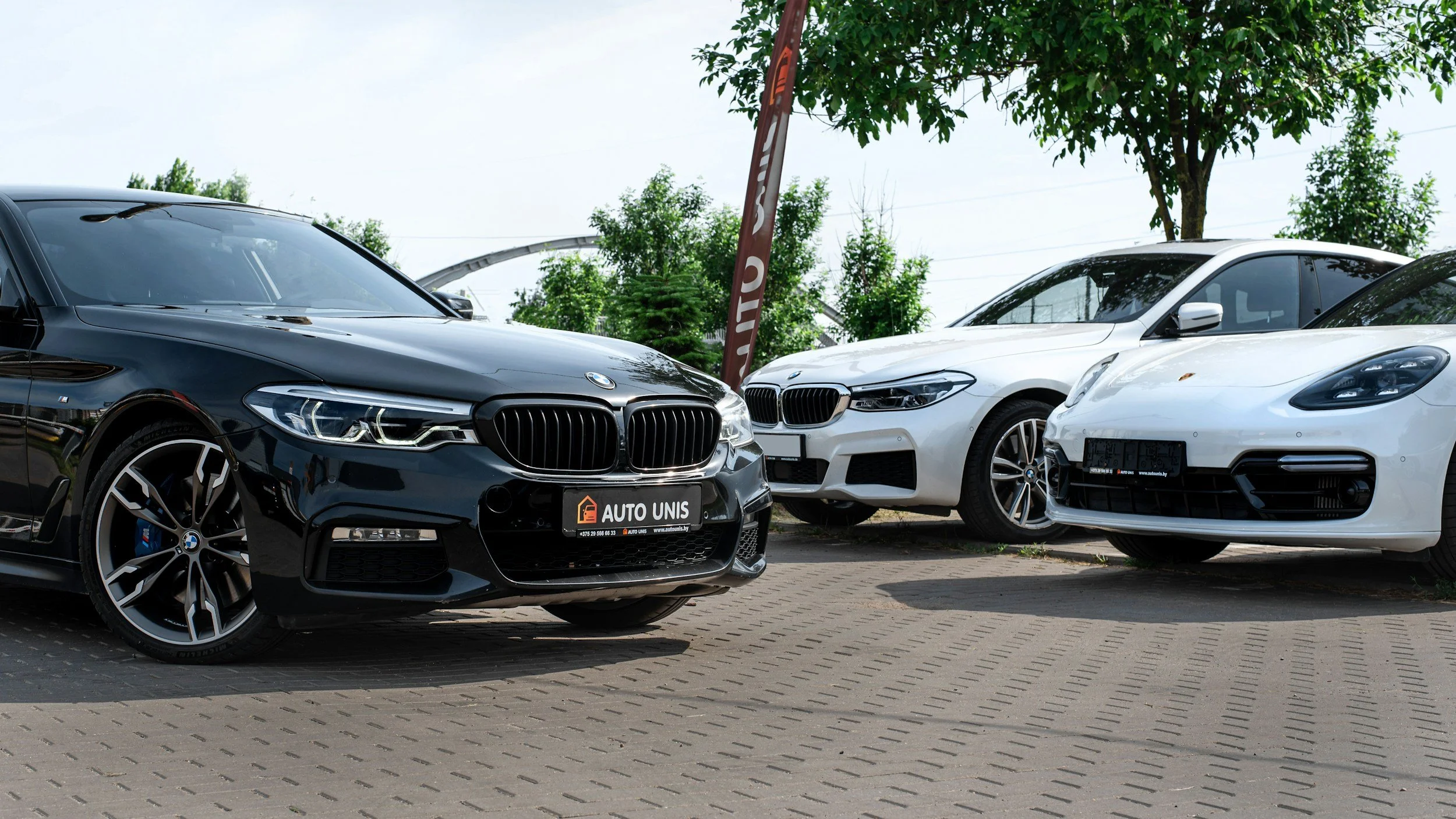 Three cars parked outside, including a black BMW on the left and two white BMWs on the right, with trees and a blue sky in the background.