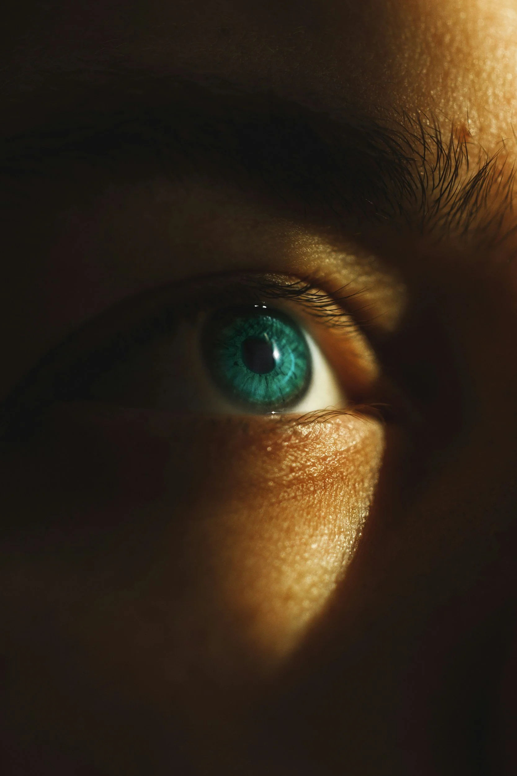 Close-up of a person's eye with a bright turquoise iris, illuminated by warm light highlighting the skin texture.