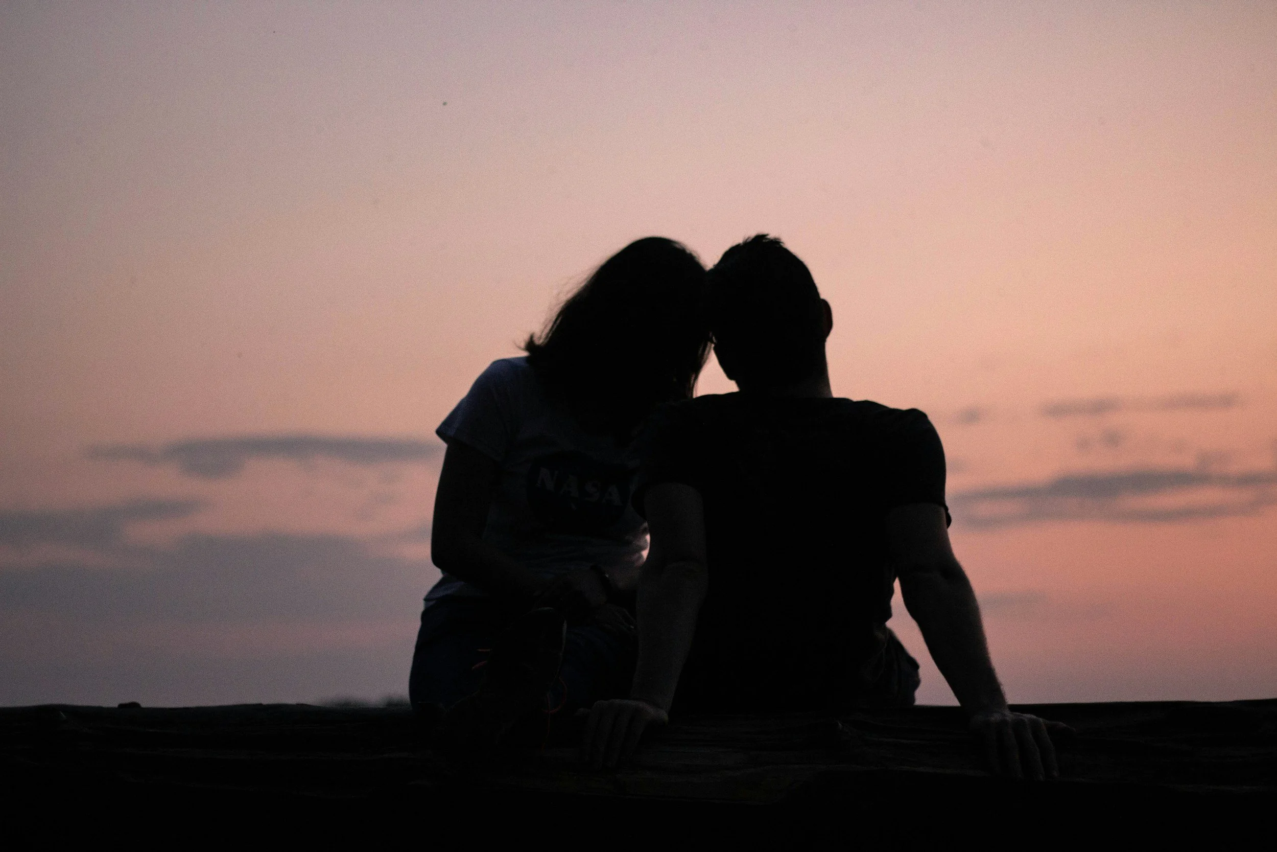 A silhouette of a couple sitting close together on a surface during sunset or dusk, with the sky painted in soft pink and purple hues in the background.