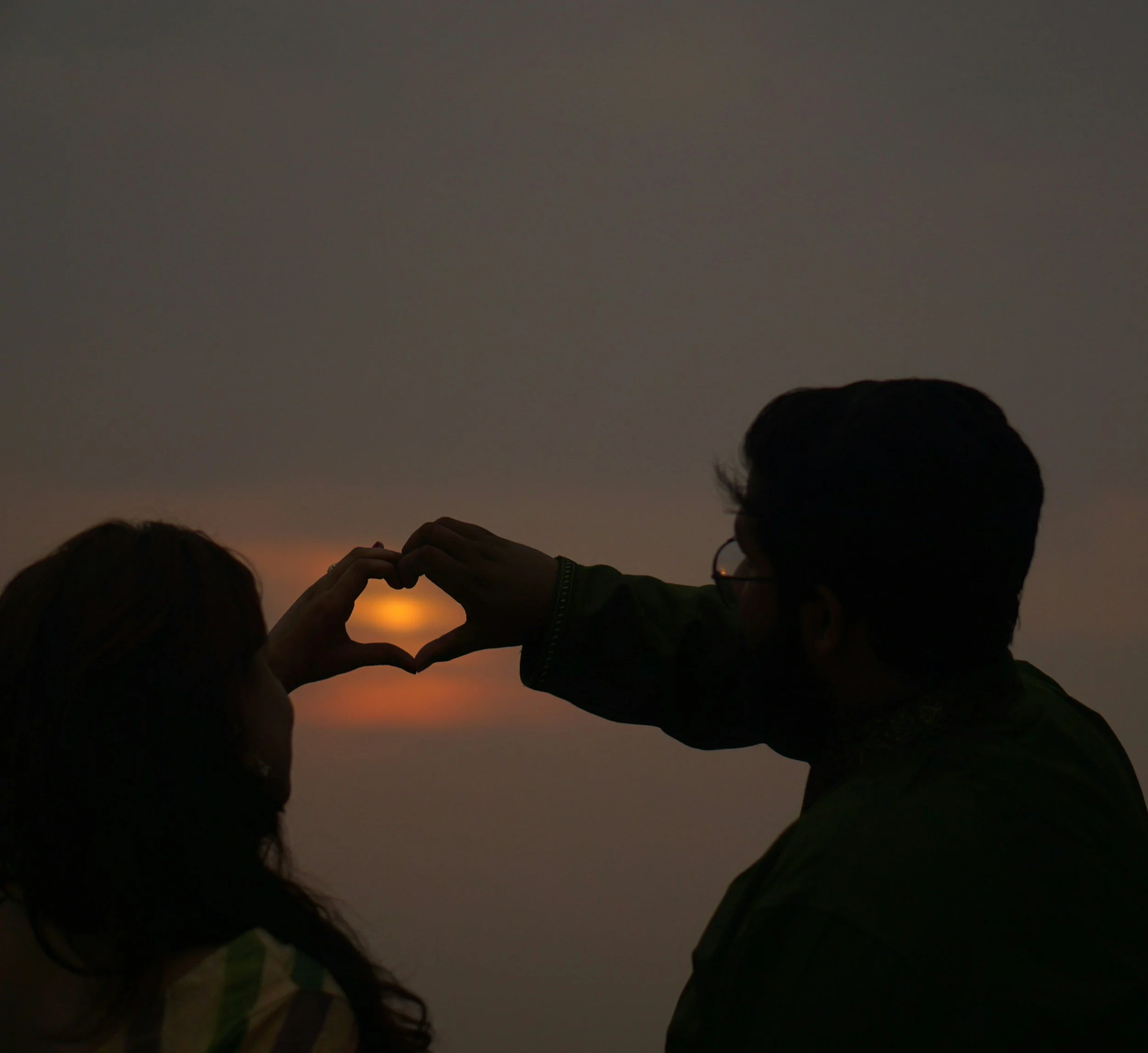 Silhouettes of a man and woman forming a heart shape with their hands around the setting sun during sunset or sunrise.