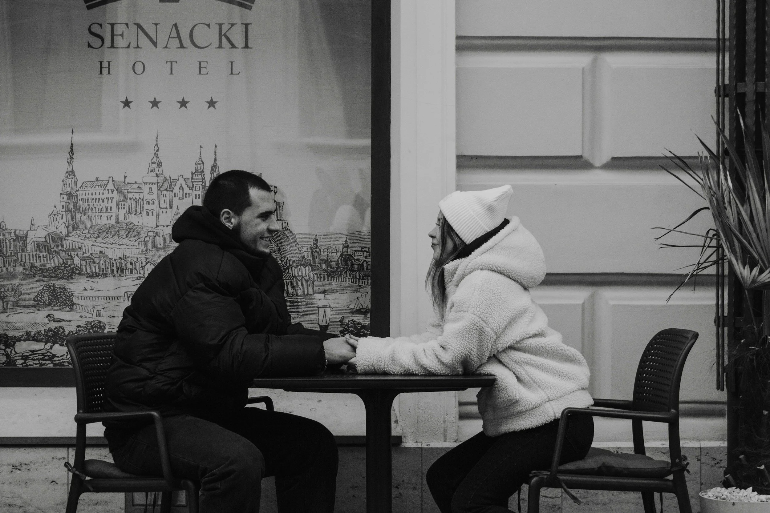 A man and a woman are sitting at a small table outside a cafe, holding hands and smiling at each other. The woman is wearing a white beanie and a fluffy jacket, while the man is wearing a dark hoodie. There is a sign in the background that reads 'SENACKI HOTEL'.