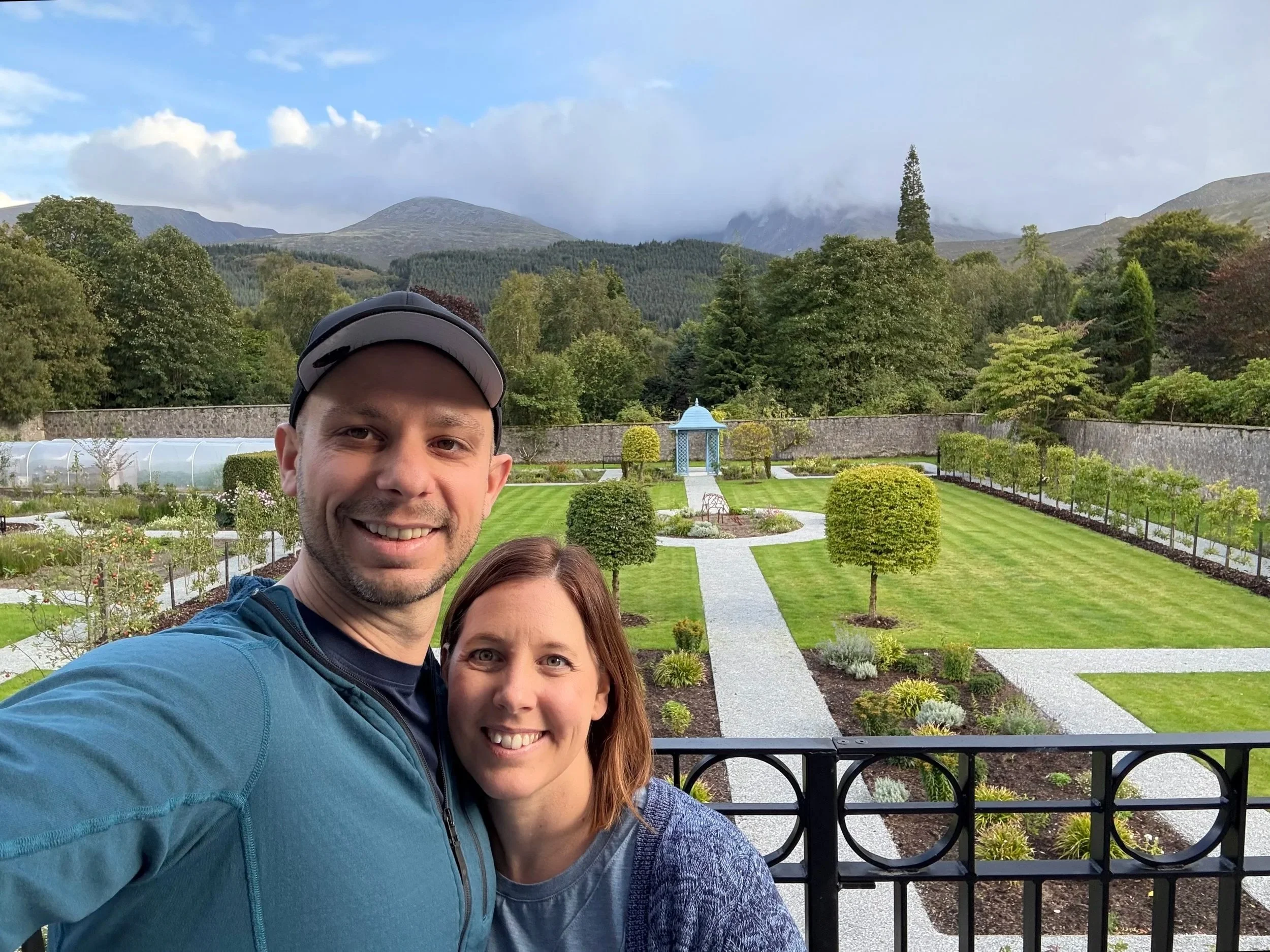 A smiling man and woman taking a selfie outdoors in front of a landscaped garden with a gazebo, trees, mountains, and cloudy sky in the background.