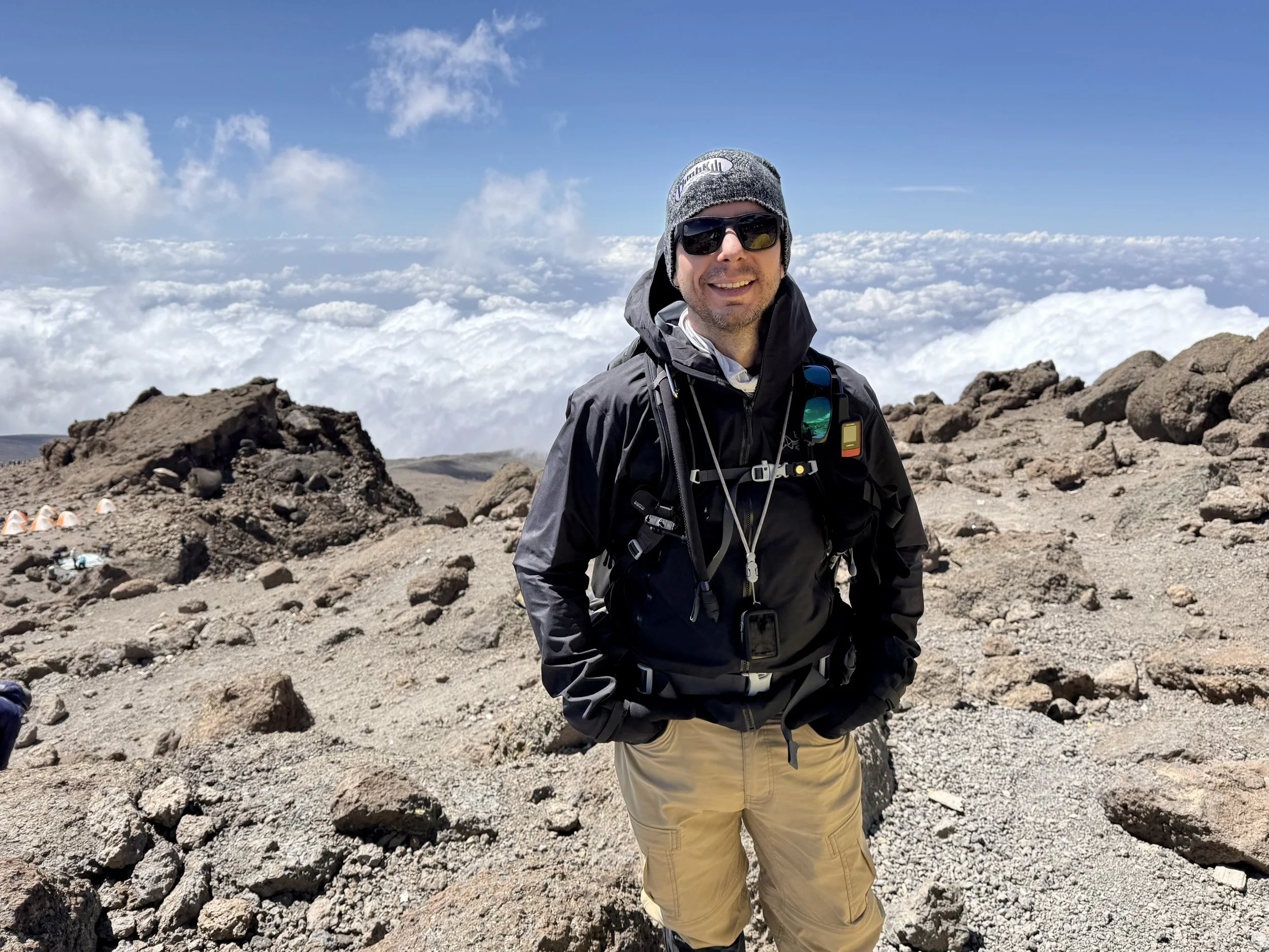 Man in outdoor gear standing on rocky mountain terrain with clouds and blue sky in the background.