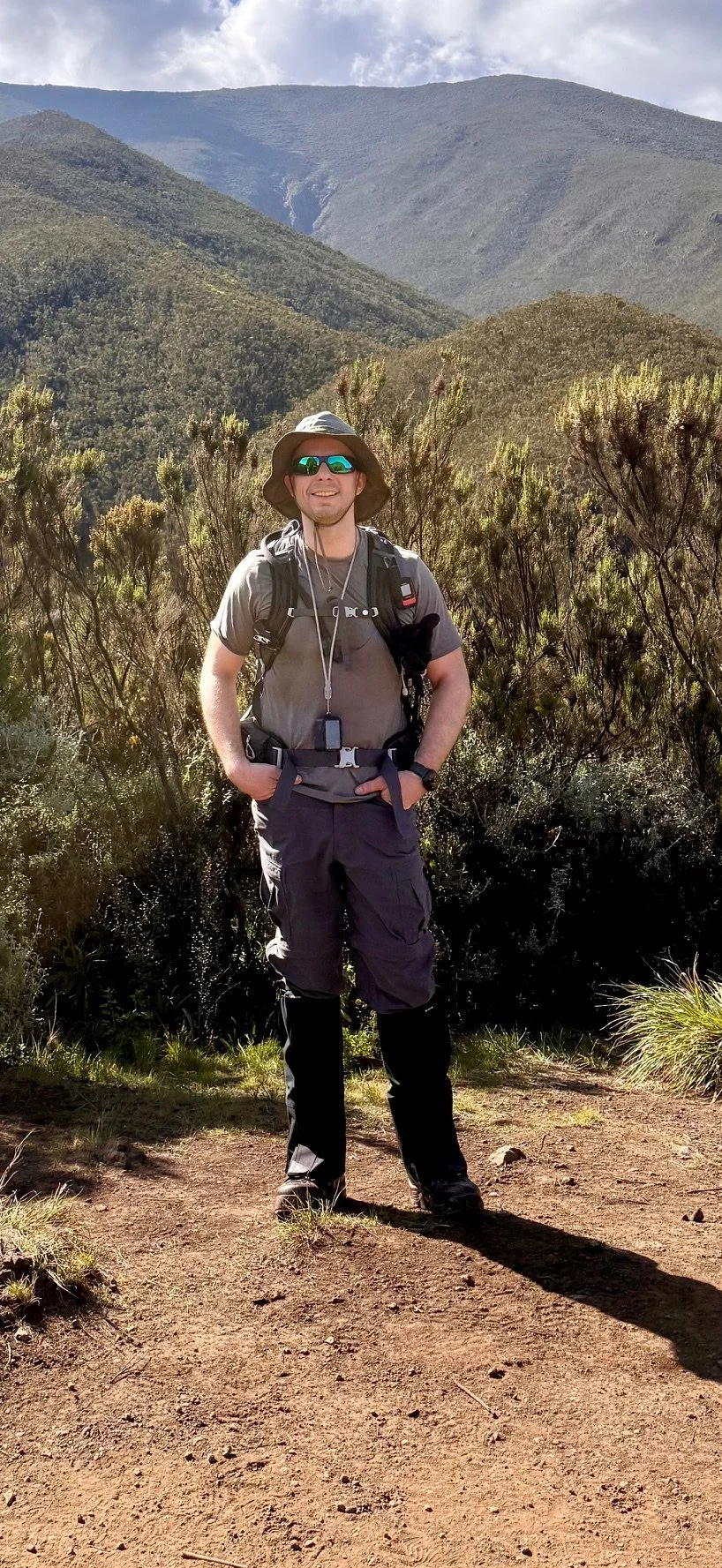 A man in outdoor hiking gear standing on a dirt trail in a mountainous landscape with hills and cloudy sky in the background.