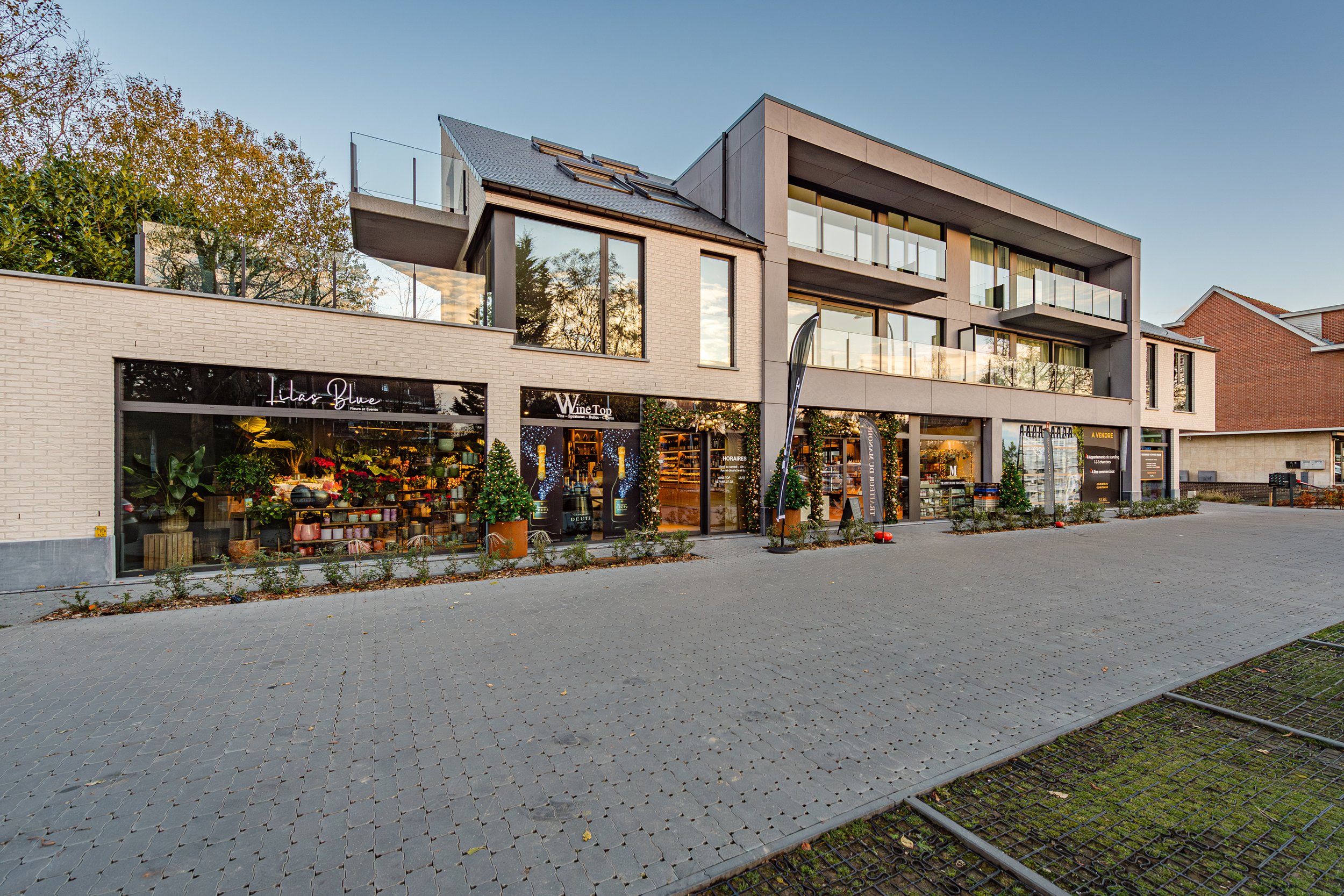 Un bâtiment moderne avec des boutiques au rez-de-chaussée, des balcons en verre au premier étage, et un espace piétonnier pavé devant. La façade est en pierre claire avec une décoration de Noël visible dans les vitrines.