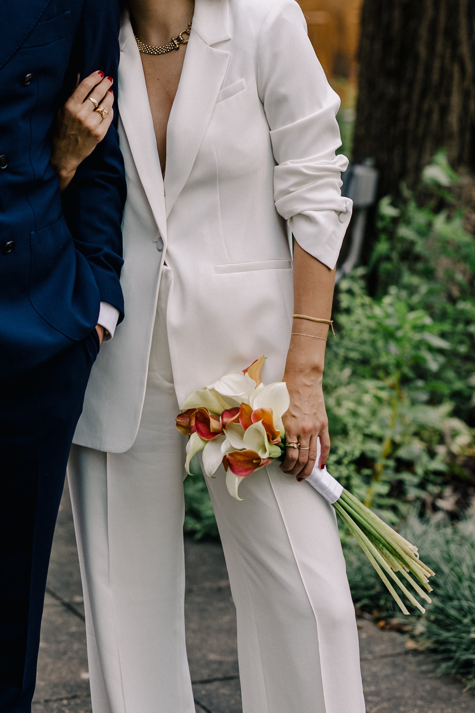 Person dressed in a white suit holding a bouquet of white and reddish-orange calla lilies, standing next to someone in a dark blue suit, outdoors with greenery.