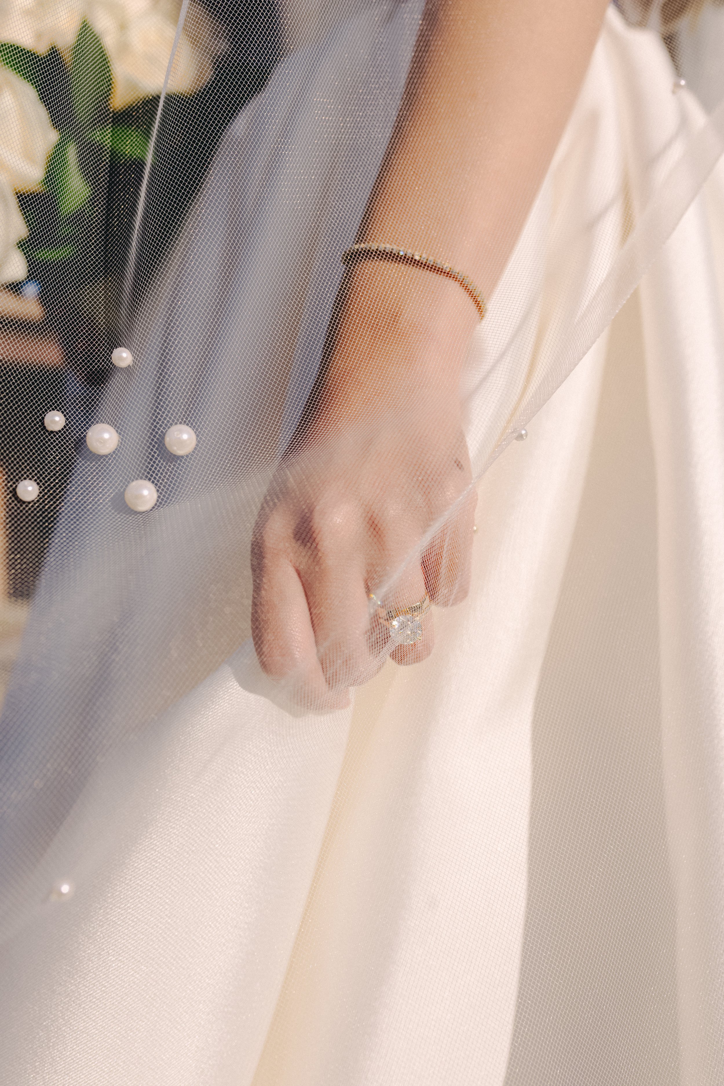 Close-up of a woman's hand and part of her dress, with veil, pearl bracelet, and ring, holding a bouquet of white roses and greenery.