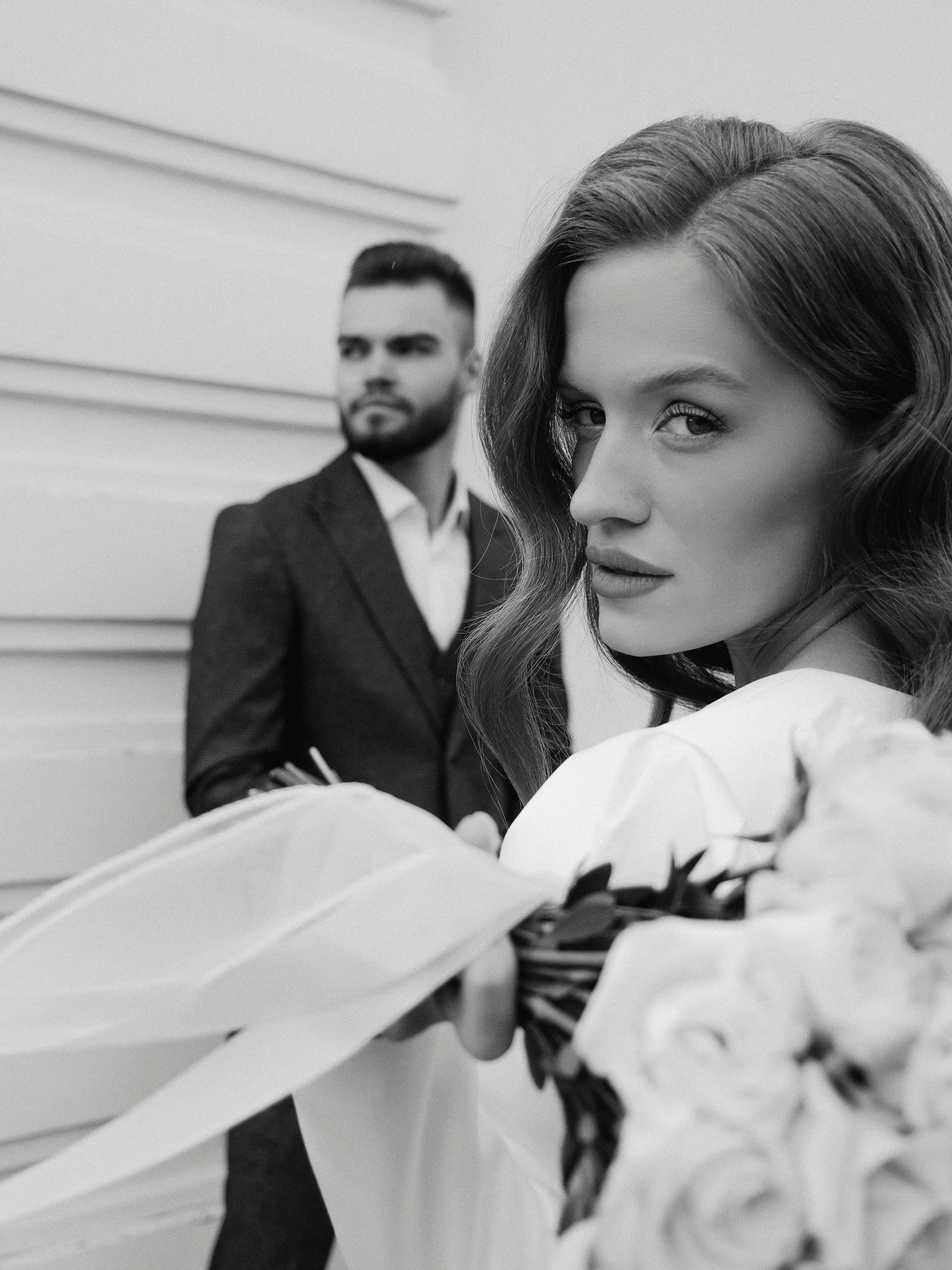 A black and white photo of a woman holding a bouquet of flowers in the foreground, with a man in a suit standing in the background, both looking in different directions.