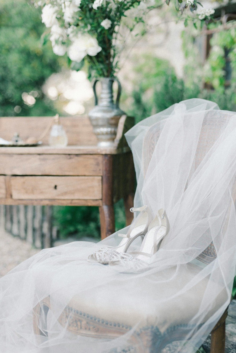 White high-heeled shoes placed on a chair draped with a white veil, outdoors with a wooden table decorated with a silver vase of white flowers in the background.