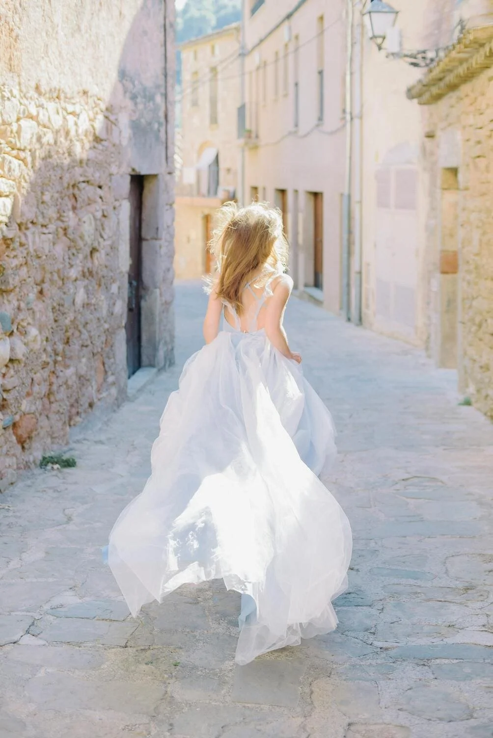 A young girl in a white dress walking down a narrow stone alleyway with old buildings on both sides, illuminated by sunlight.
