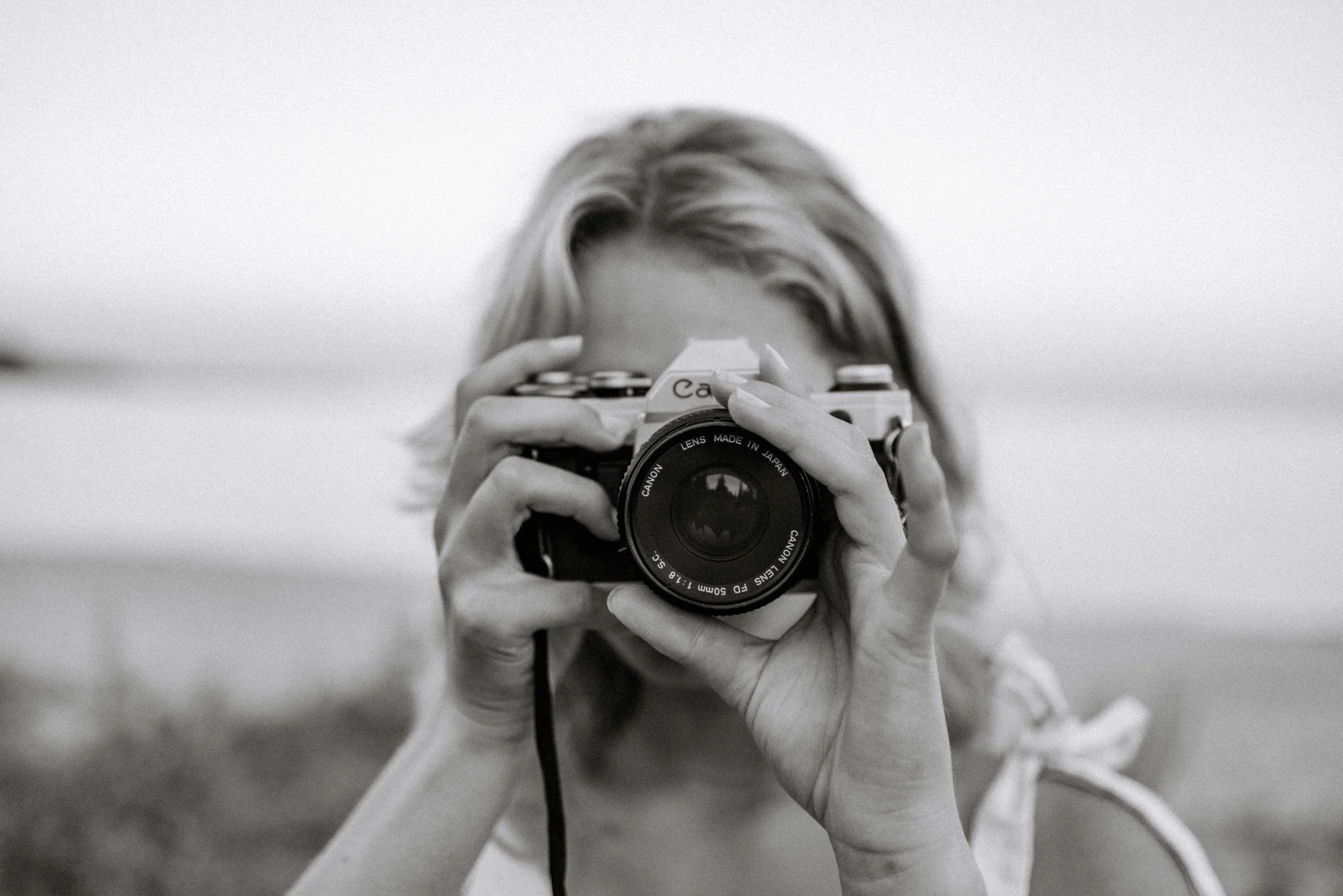 A woman holding a camera up to her face, looking through the viewfinder, in black and white.