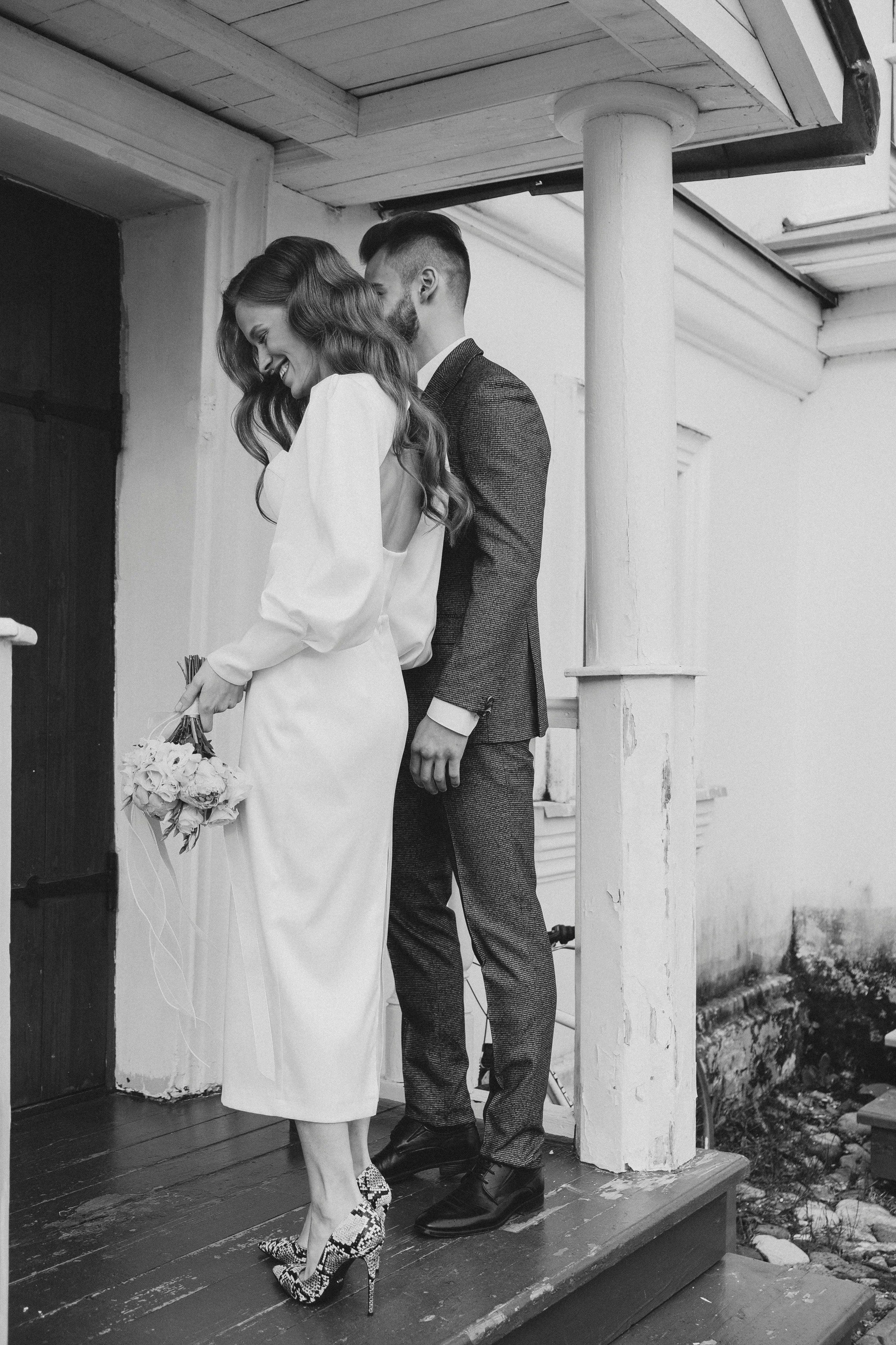 A black and white photo of a smiling woman in a white dress holding a bouquet, standing close to a man in a suit on a small porch, both smiling and leaning towards each other.