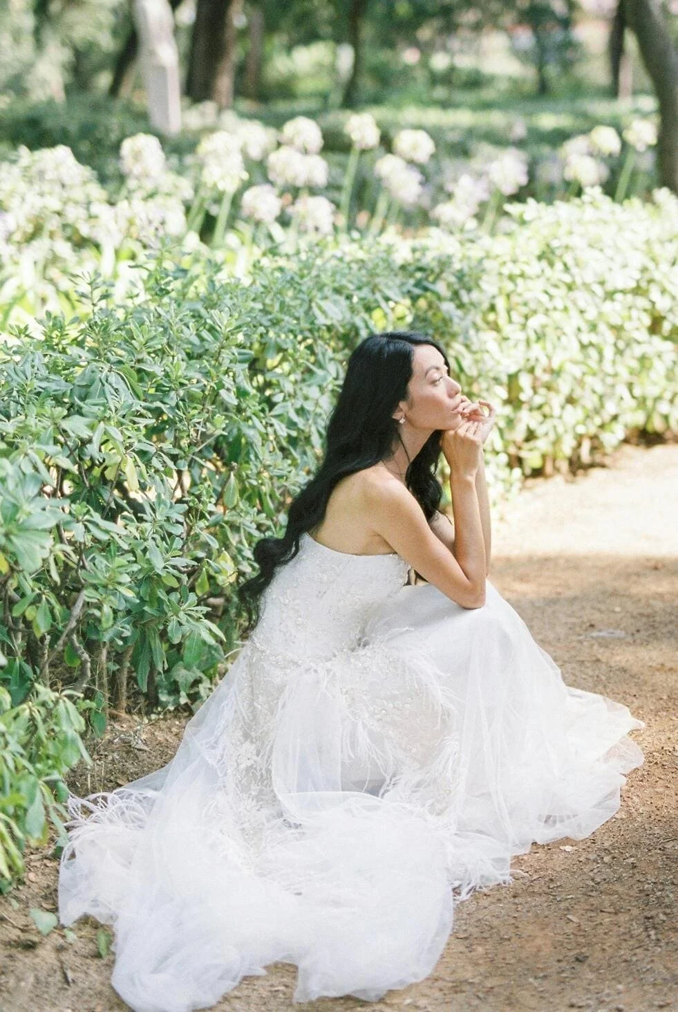 Woman in a white dress sitting on the ground next to green bushes in a garden or park.