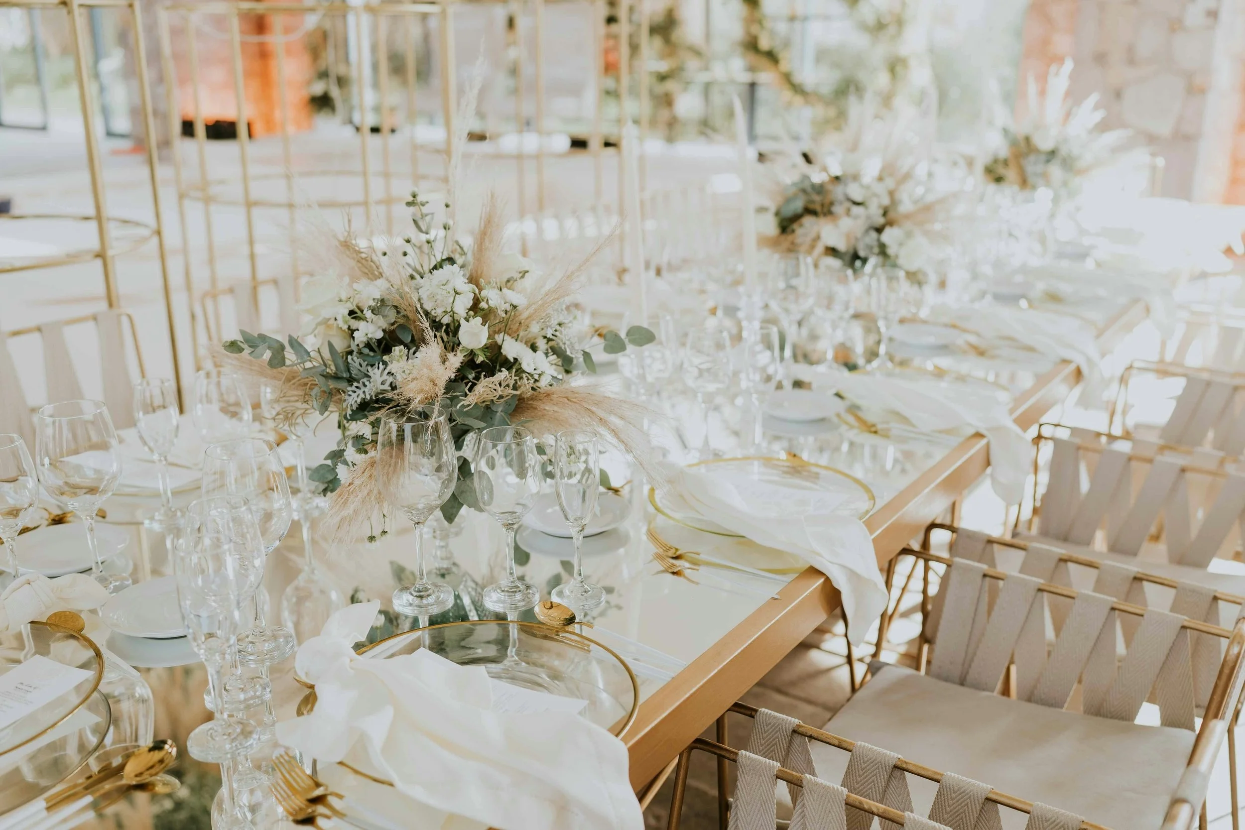 Elegant banquet table set with white plates, gold utensils, glassware, and white cloth napkins, decorated with floral centerpieces featuring white flowers and greenery, in a bright room with large windows and brick walls.