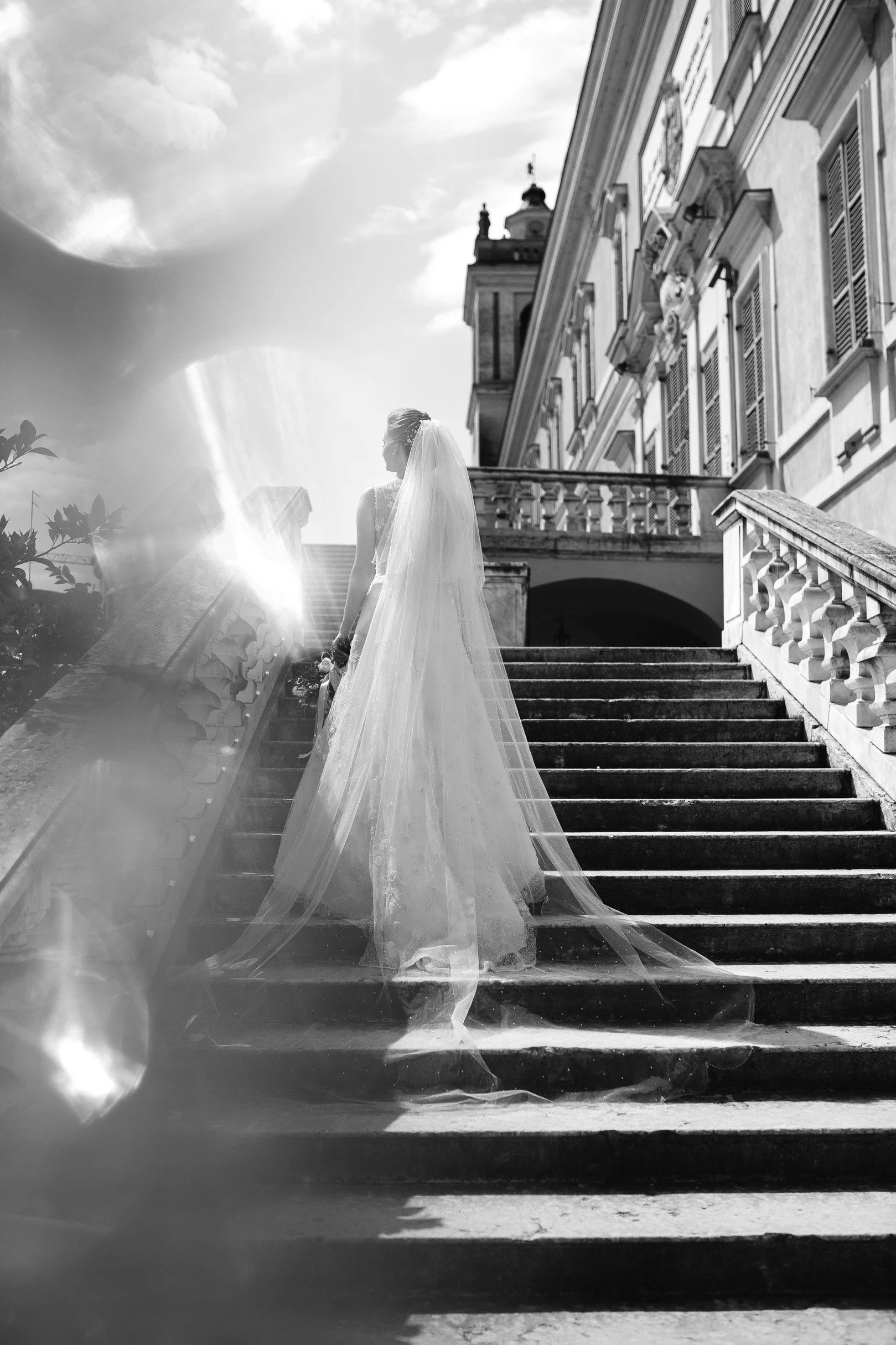 Bride in a wedding gown with veil, ascending outdoor stone stairs in front of a large historic building, with dramatic sunlight and clouds in the sky.