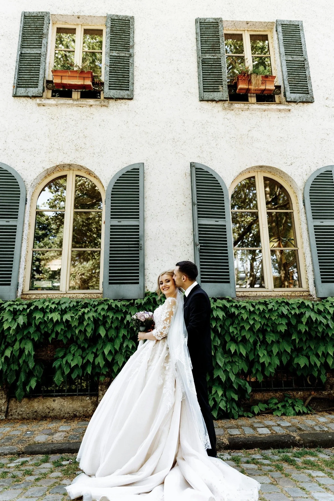 A bride in a white wedding gown holding a bouquet of flowers, and a groom in a black suit, standing close together and smiling in front of a green ivy-covered wall with large arched windows and blue shutters, in front of an old white building.