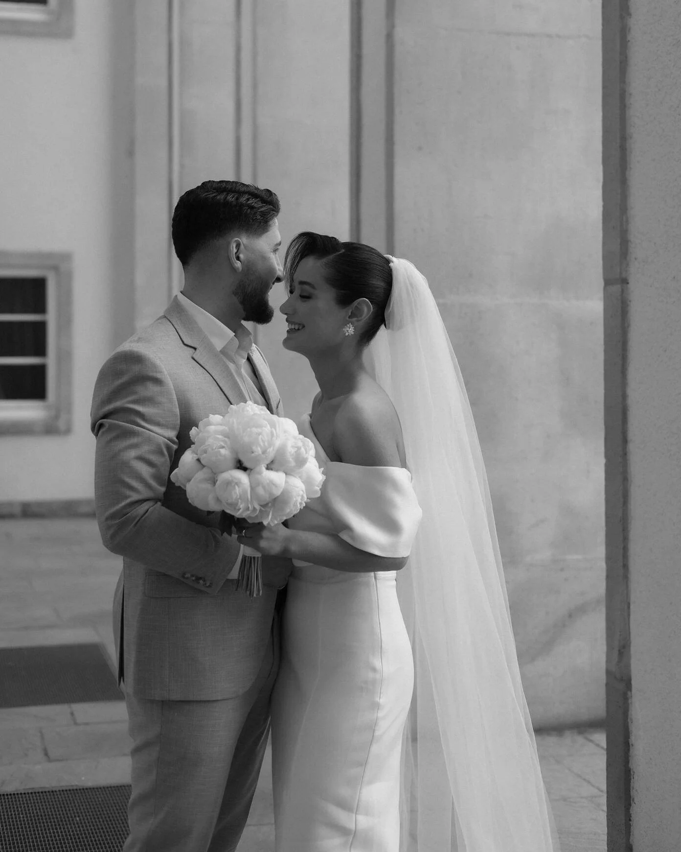 A black and white photo of a bride and groom standing close, smiling, with the bride holding a bouquet of flowers, in an indoor setting.