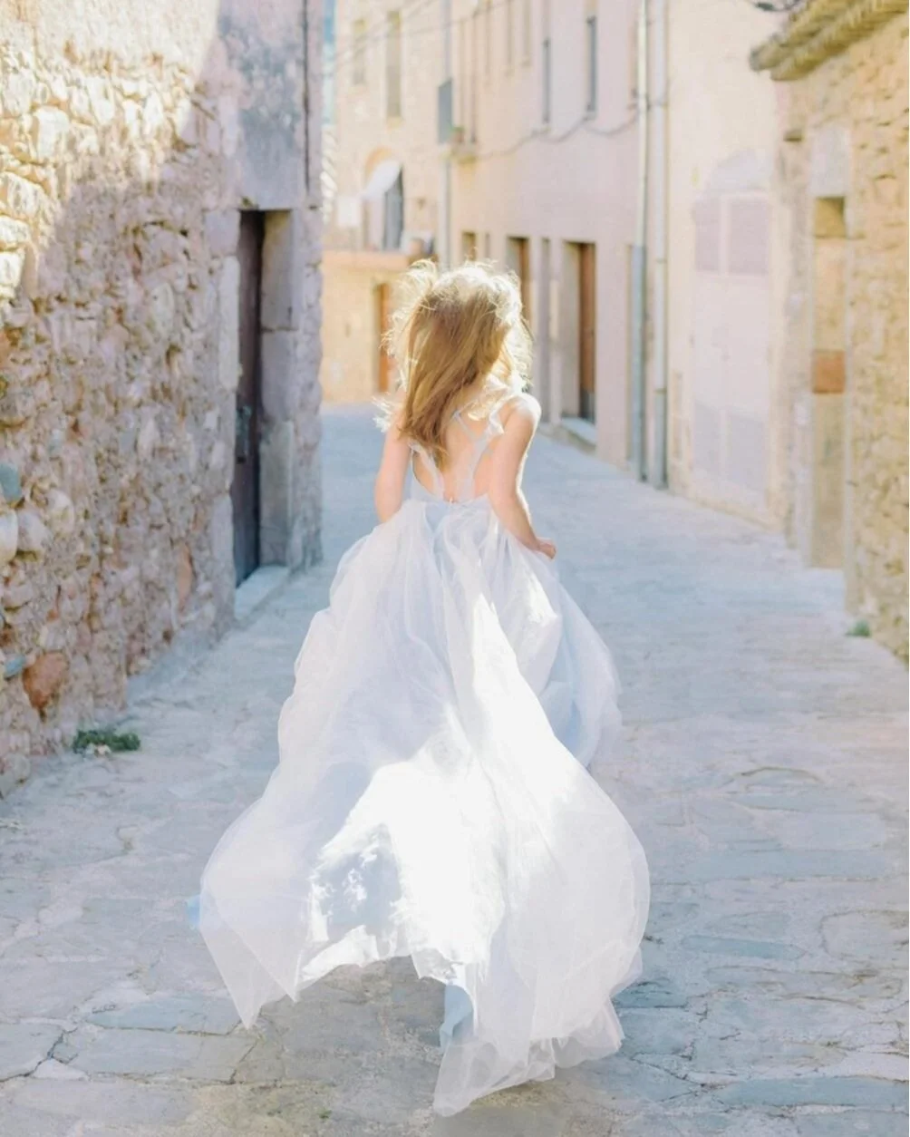 A young girl in a white dress walking down a narrow stone alleyway with old stone and brick buildings on either side, sunlight illuminating her hair and dress.
