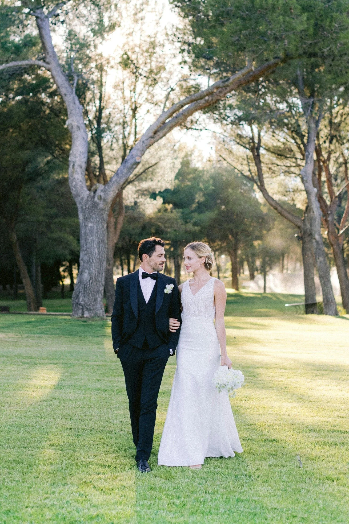 A bride and groom walking arm in arm on a grassy field in a park with tall trees and sunlight filtering through the leaves.