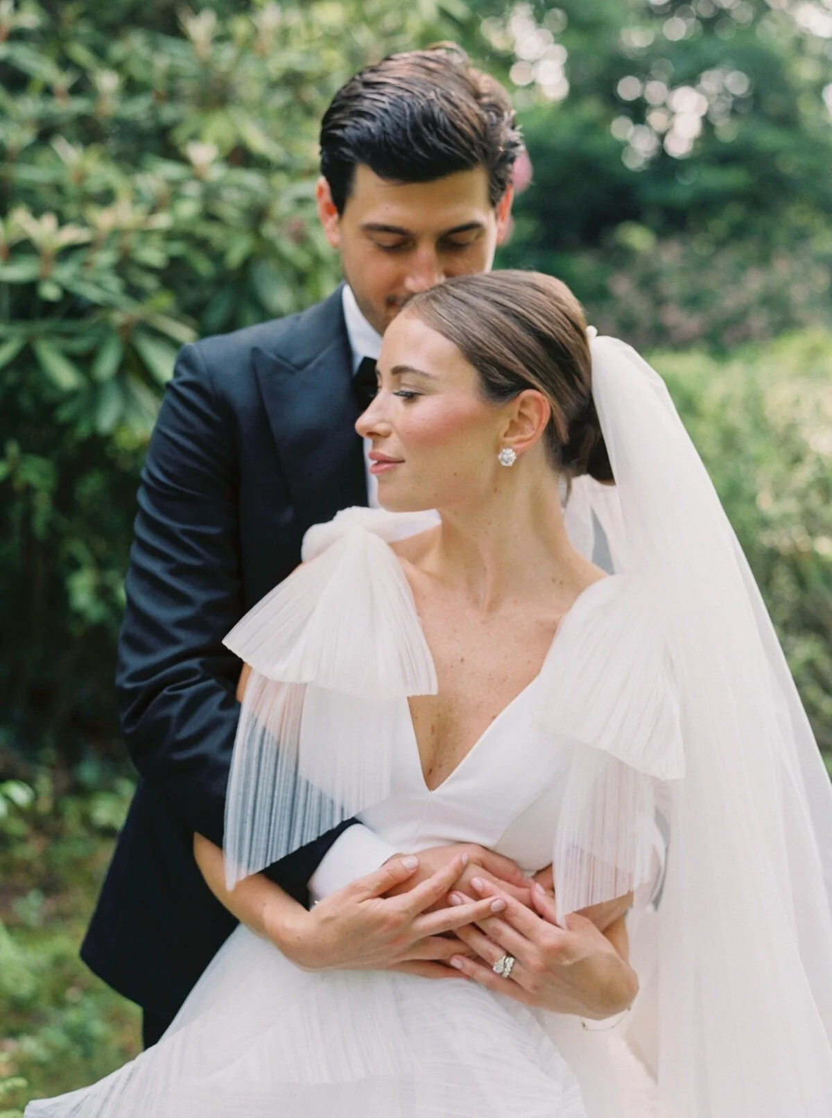 A bride and groom embracing outdoors, with the groom standing behind the bride, both looking content and tender. The bride is wearing a white dress with pleated details and a veil, and the groom is in a dark suit. The background features lush green foliage.