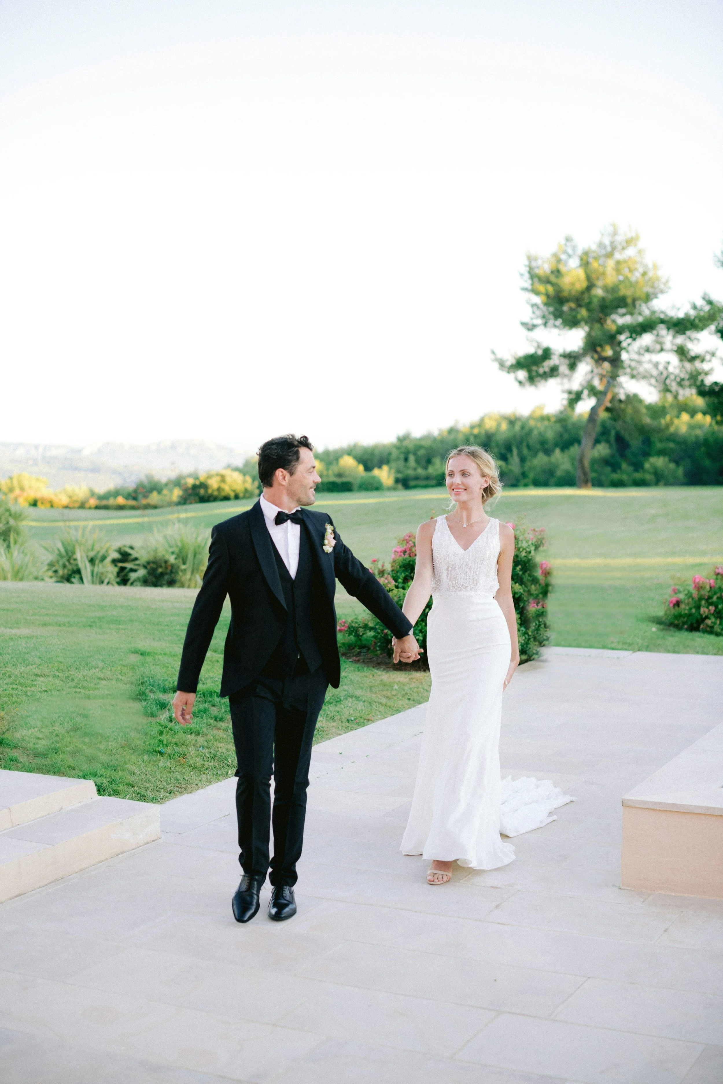 A bride and groom walking hand in hand outdoors on a bright day, surrounded by greenery and pink flowering bushes.