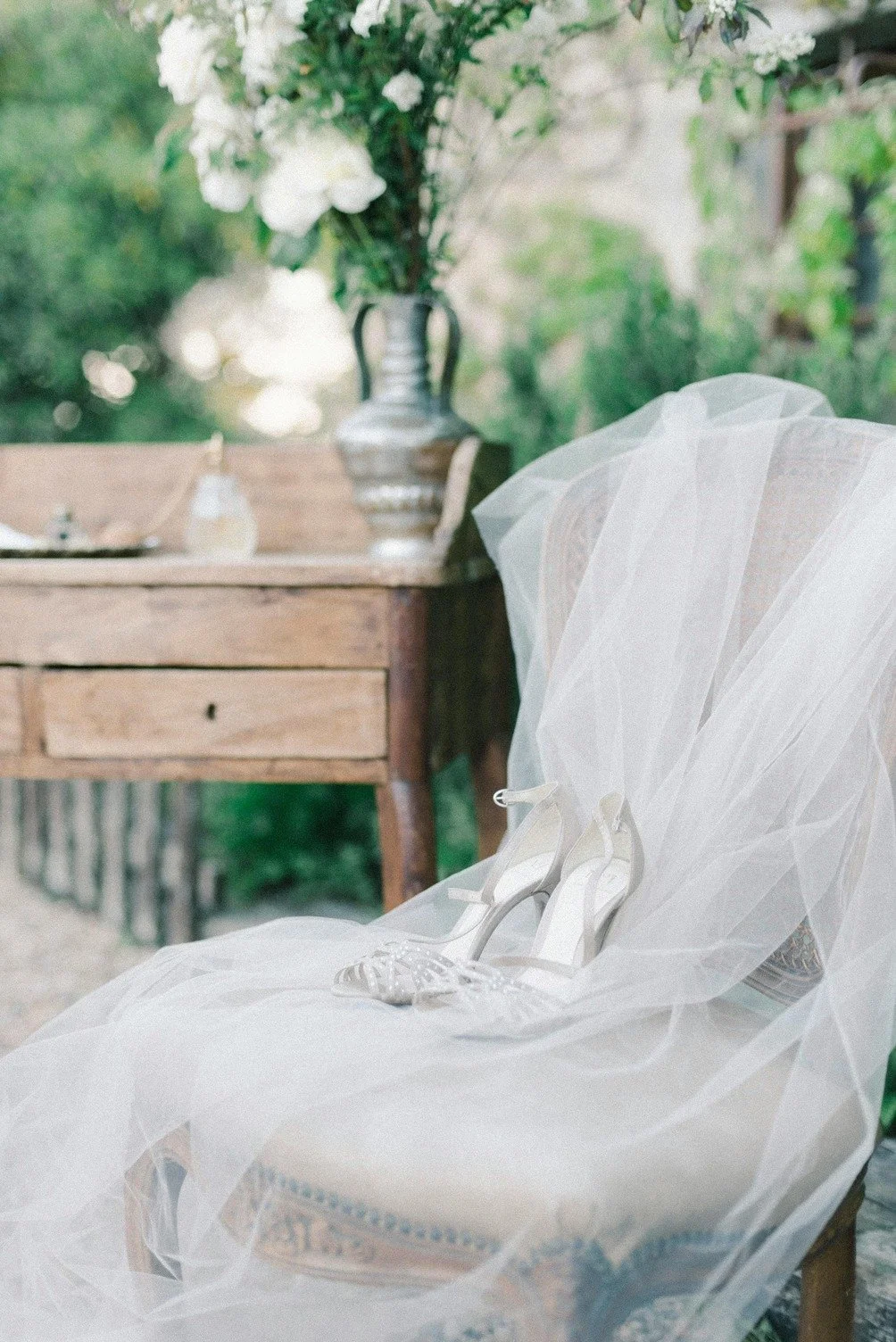 White wedding high heels placed on a chair with a white veil draped over it, outdoors with a wooden table and a large flower arrangement in the background.
