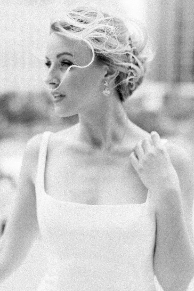 A black-and-white photo of a woman with short, wavy blonde hair, wearing earrings and a white sleeveless top, looking to her left with a gentle smile.