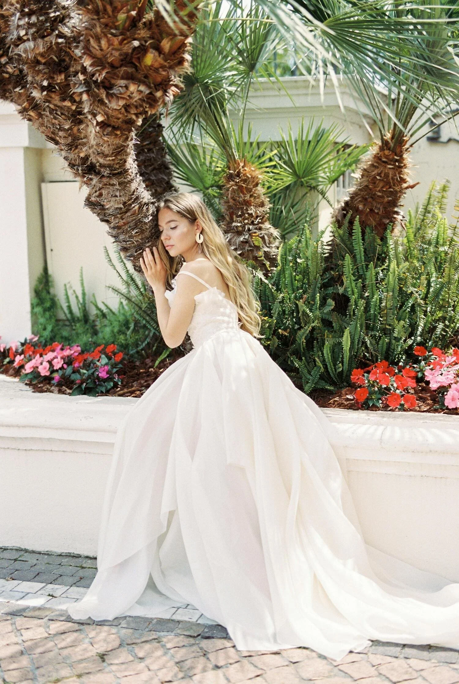 A young woman in a white elegant dress with long flowing skirt, posing beside a palm tree and lush green plants with colorful flowers in an outdoor garden setting.