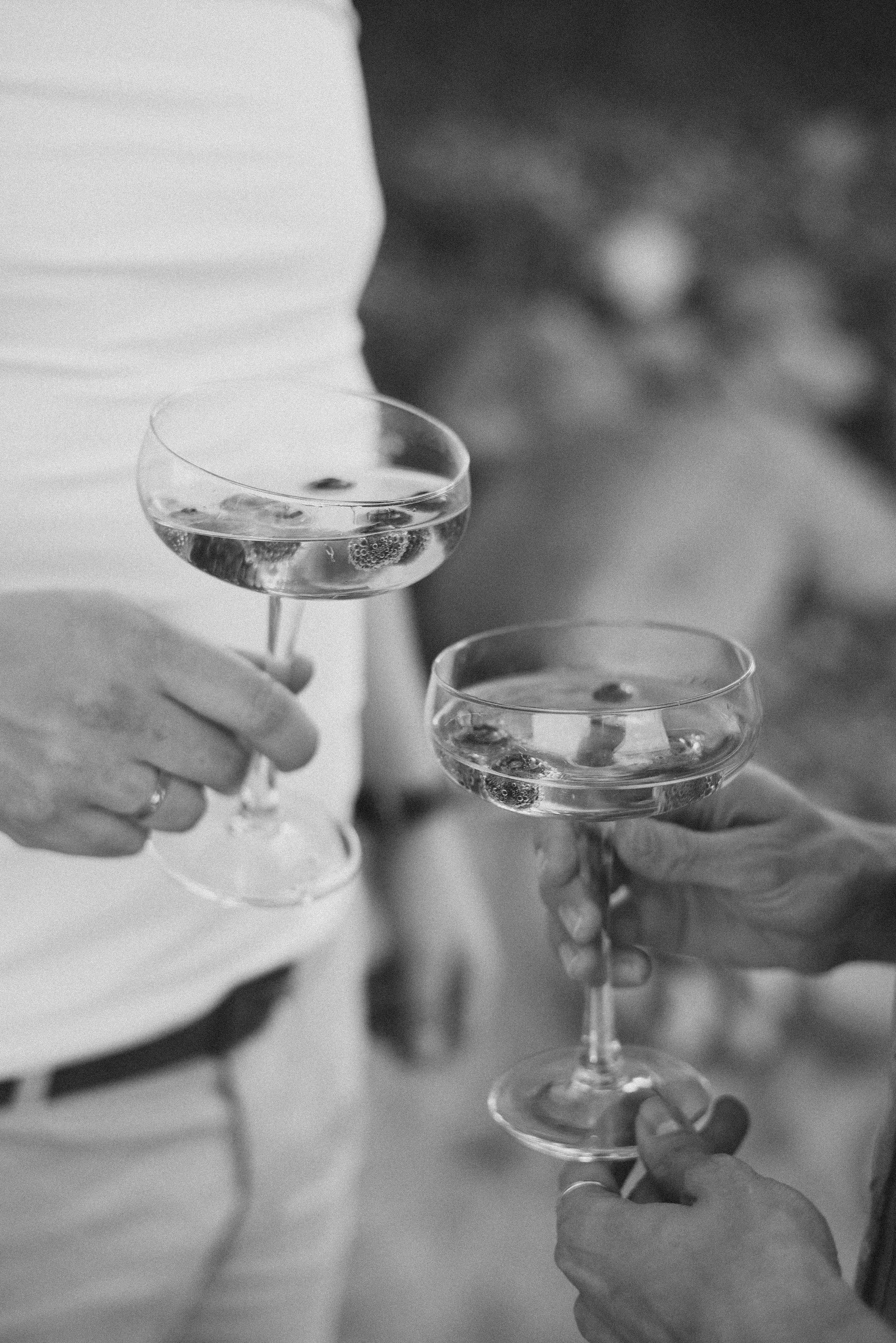Two people holding coupe glasses filled with a clear beverage, possibly champagne or cocktails, in a black-and-white photo.