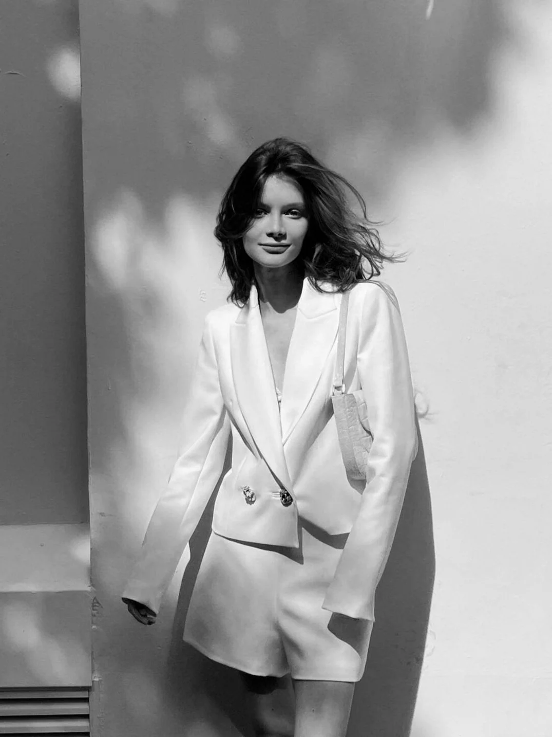 Black and white photo of a woman with shoulder-length wavy hair, wearing a tailored blazer and matching shorts, standing against a plain wall with shadows. She has a confident expression and a small purse on her shoulder.