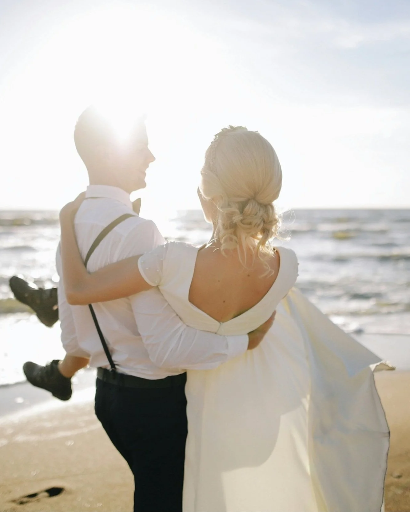 A couple in wedding attire on the beach, with the man holding the woman in his arms as they look at each other, backlit by the sun.