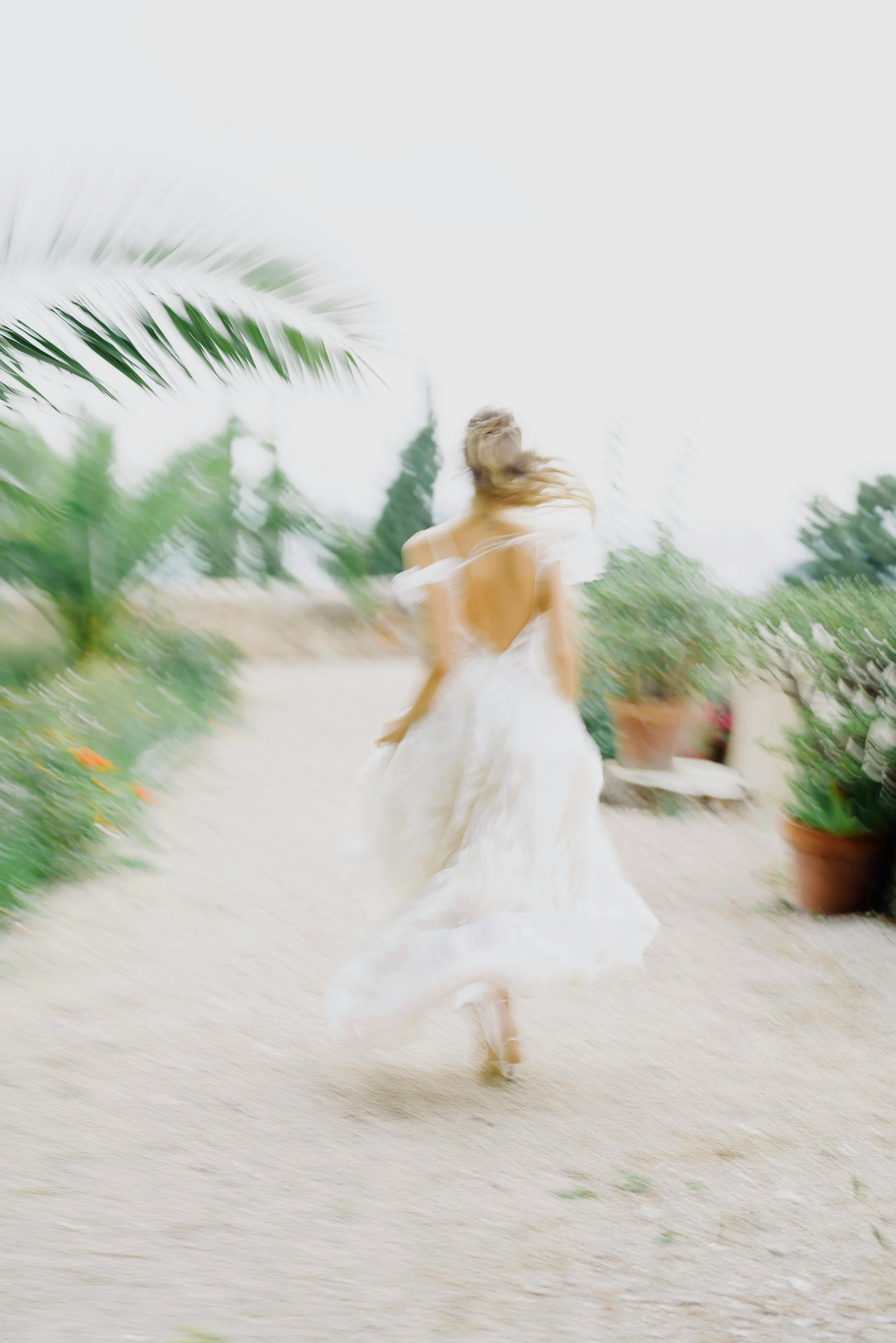 A woman in a flowing white dress running outside on a gravel path surrounded by potted plants and trees, with a blurred motion effect.