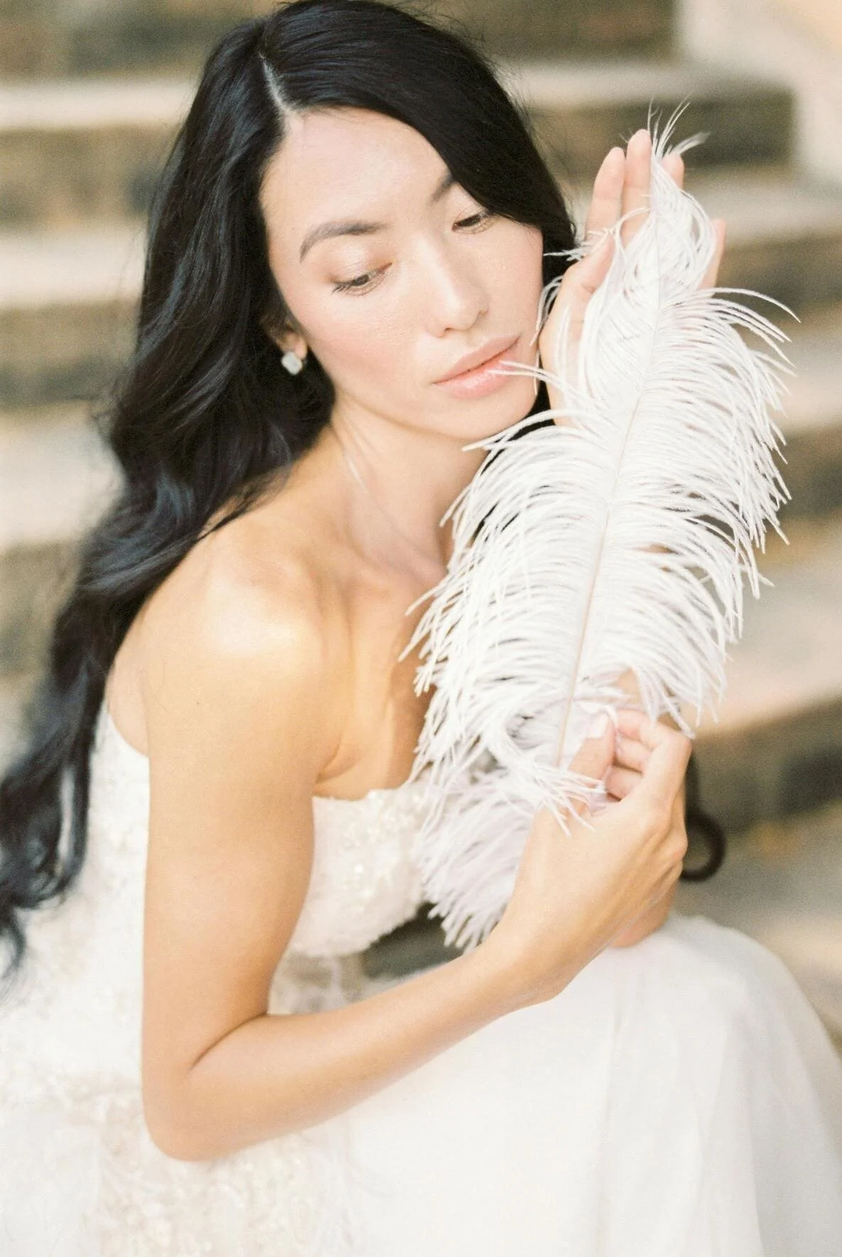 A woman with long black hair, wearing a white dress, holding a large white feather, sitting on stairs with a thoughtful expression.