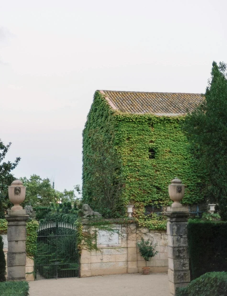 An old building covered in green ivy, with a tiled roof, flanked by stone pillars and a gate, surrounded by trees and bushes.