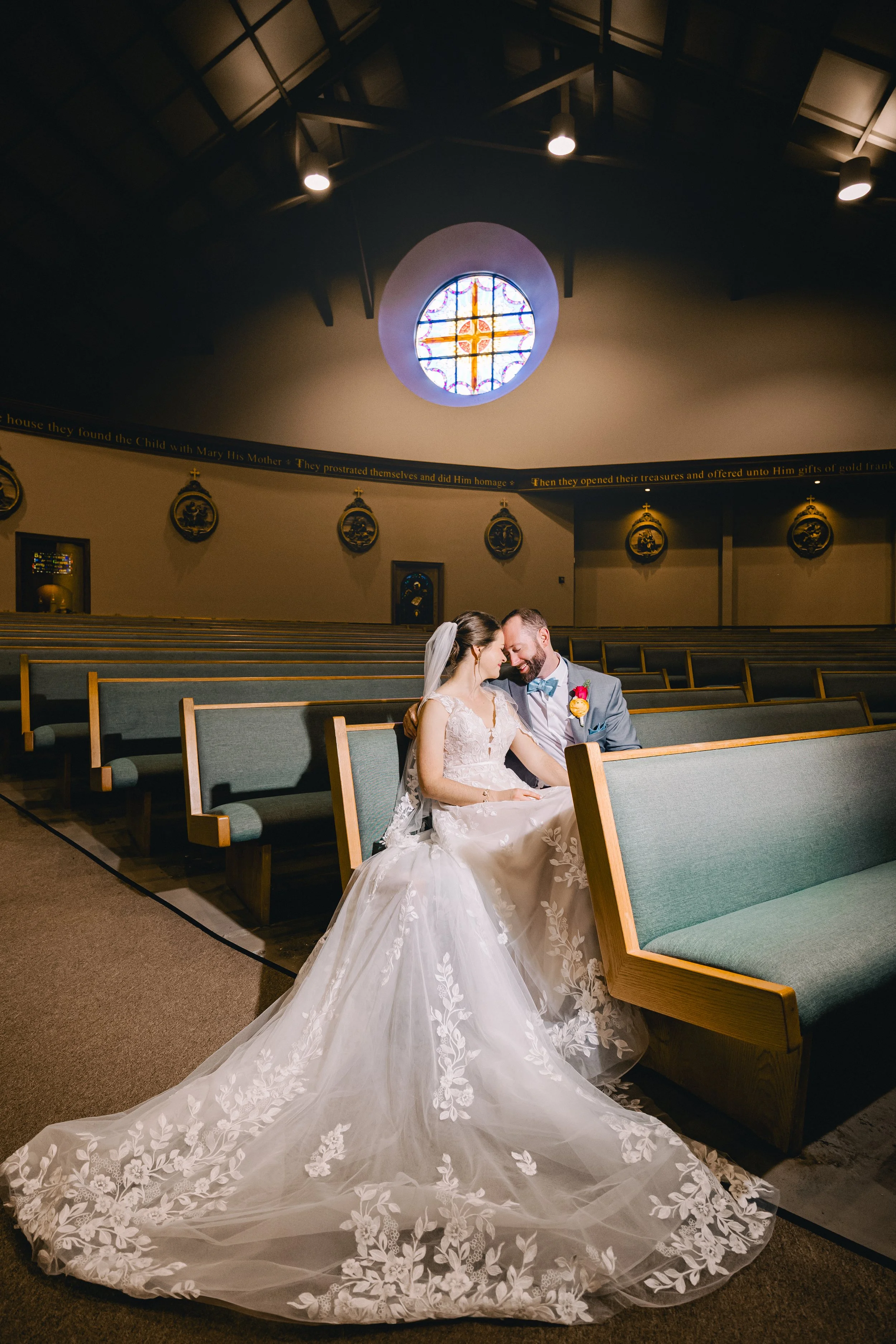 A bride and groom sitting close together on a pew in a church, sharing an intimate moment. The bride is wearing a long, white wedding gown with lace detailing and a veil, and the groom is in a gray suit with a light blue bow tie and boutonniere. The church interior features a stained glass window, religious artwork, and soft lighting.