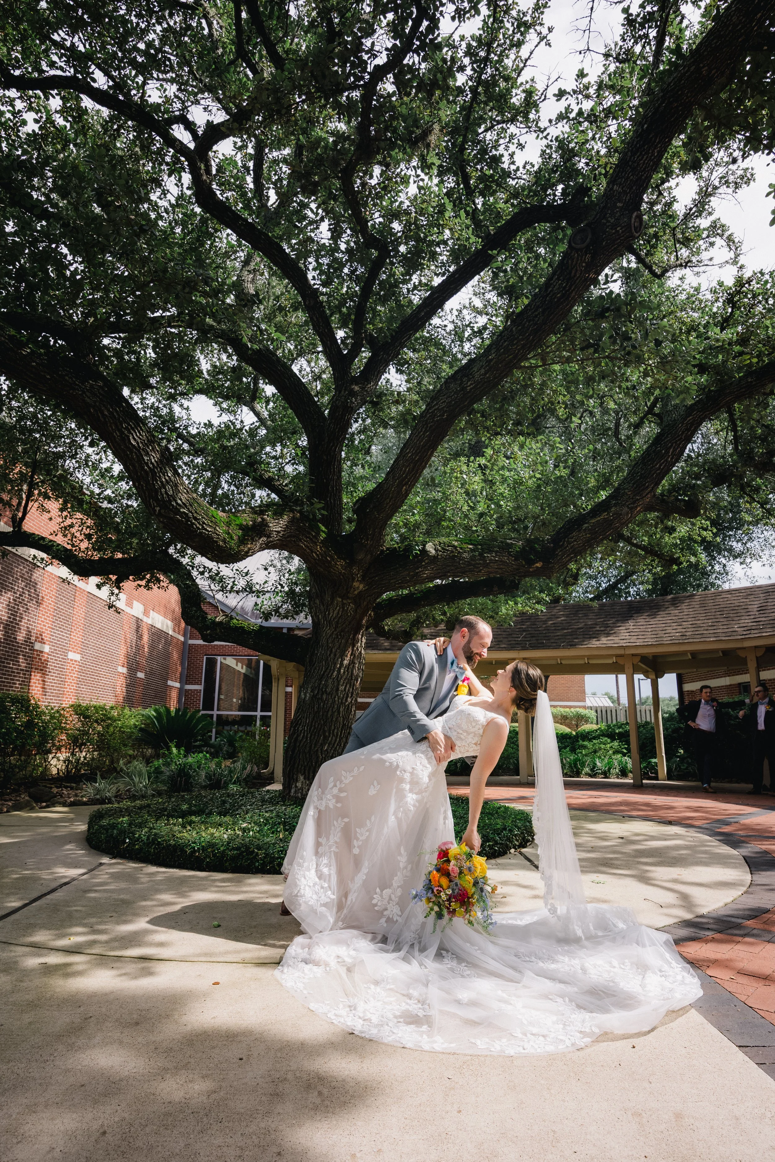 A bride and groom share a romantic dance under a large tree in an outdoor wedding setting. The bride holds a colorful bouquet, the groom is in a gray suit, and there are guests in the background.