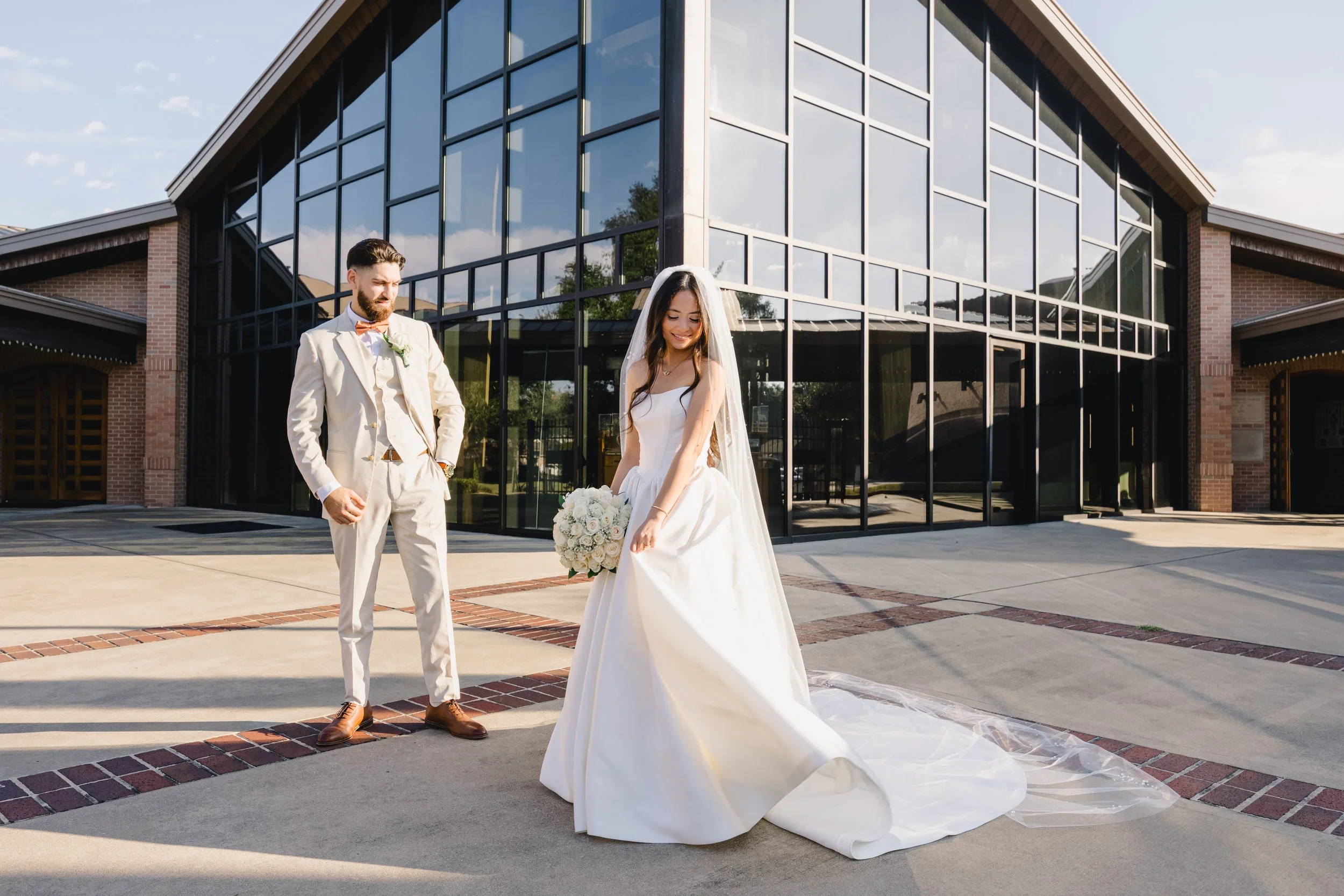 A bride in a white wedding dress holding a bouquet of white roses stands outside a modern glass building, with a groom in a cream suit and bow tie looking at her.