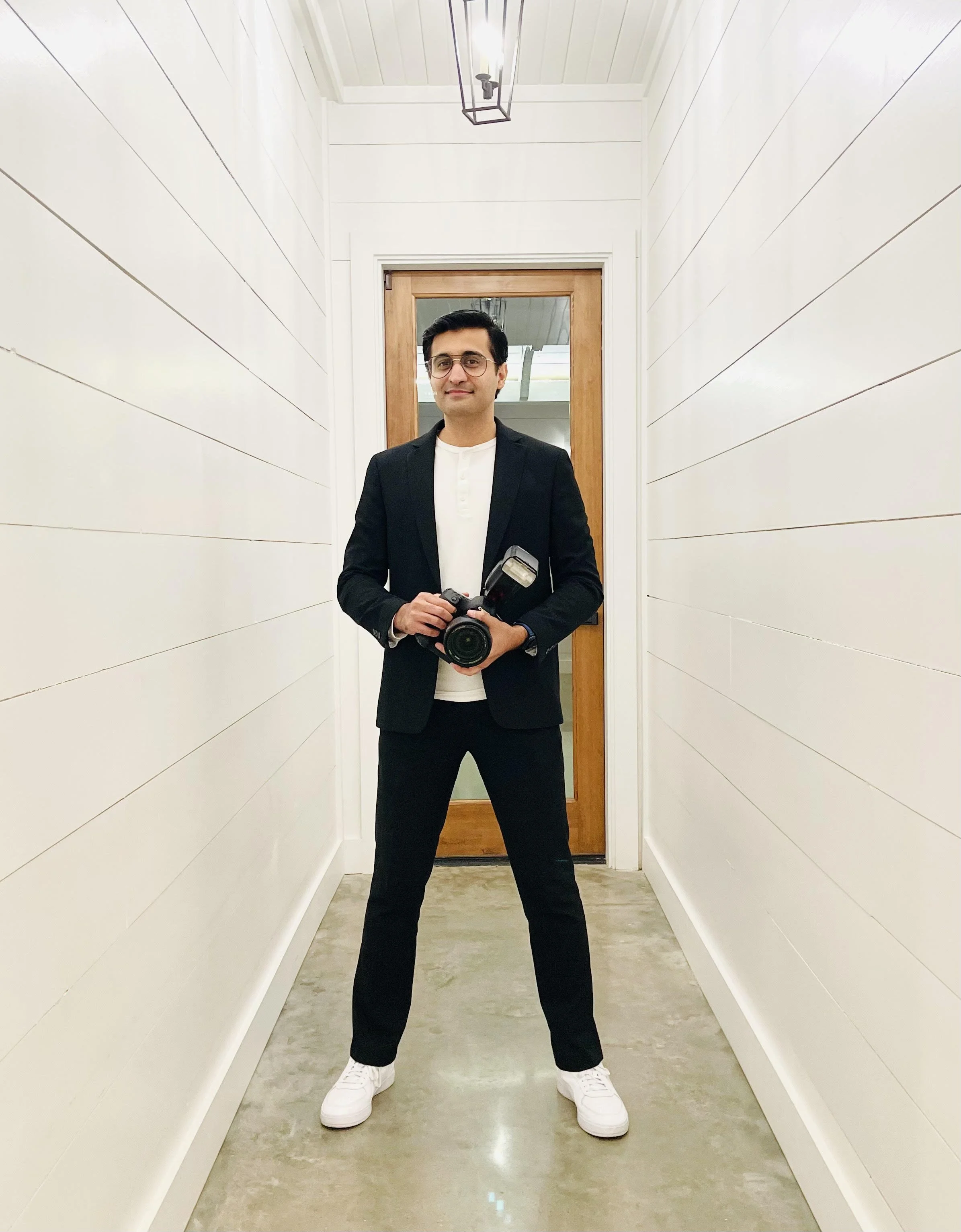 A man in a black suit with a camera and flash stands in a narrow white hallway with a wooden door behind him.